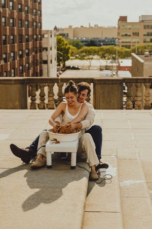 Couple sits on outdoor steps with pottery wheel sharing intimate creative moment against urban backdrop.