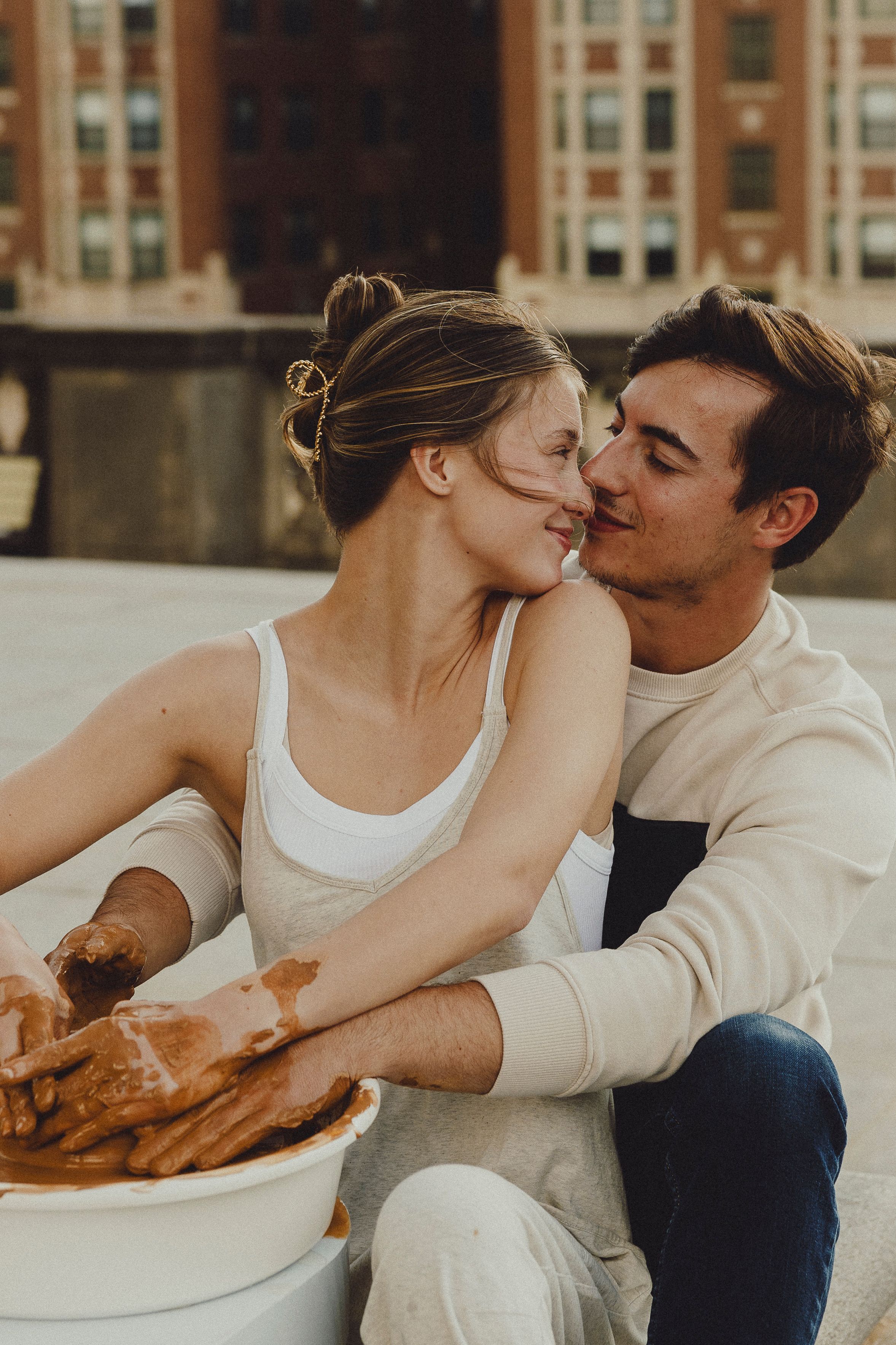 Couple kisses while working with clay at an outdoor pottery session in an urban setting.