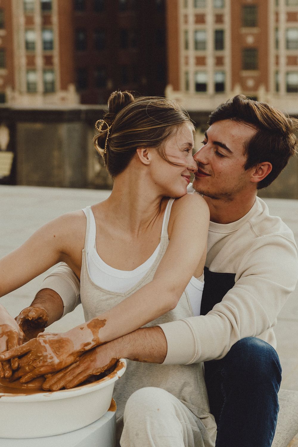 Couple kisses while working with clay at an outdoor pottery session in an urban setting.