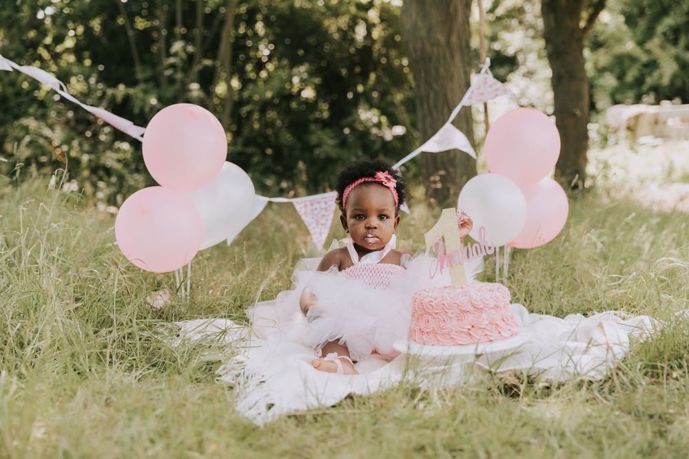 A pink birthday celebration setup in a garden with pink balloons, tulle dress, and matching cake on white blanket.
