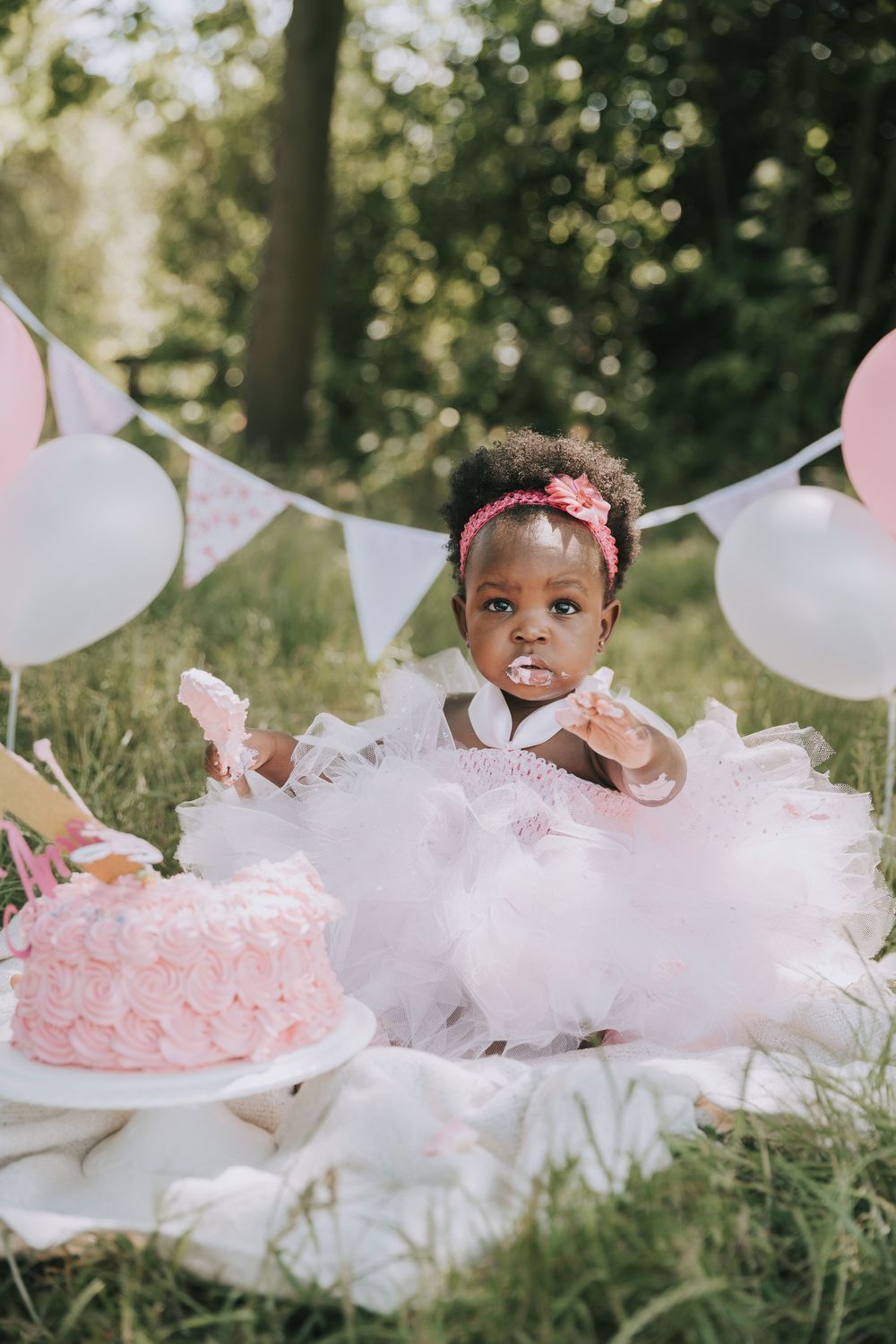 A whimsical first birthday celebration setup in a garden with pink and white decorations and a pink cake.