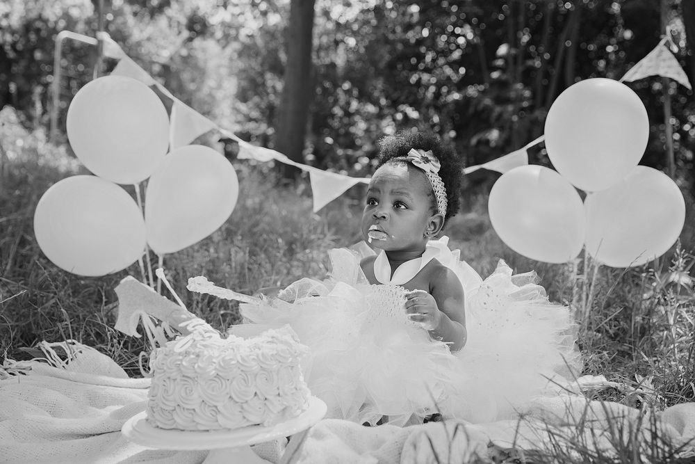 A young child in a white tulle dress sits at an outdoor birthday party with white balloons and a cake.