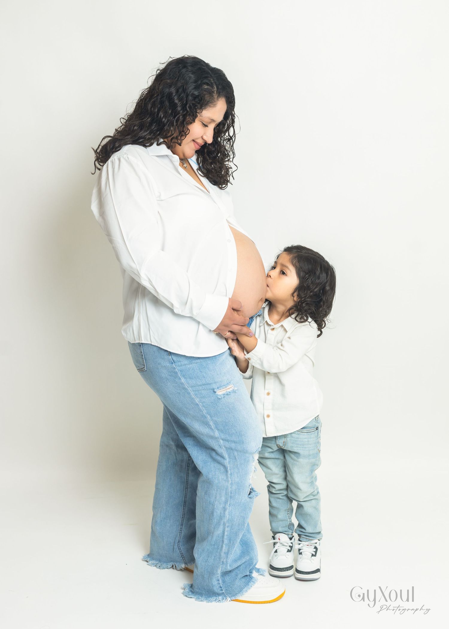 A pregnant woman in white shirt and jeans stands with a small child looking at her belly against a light background.