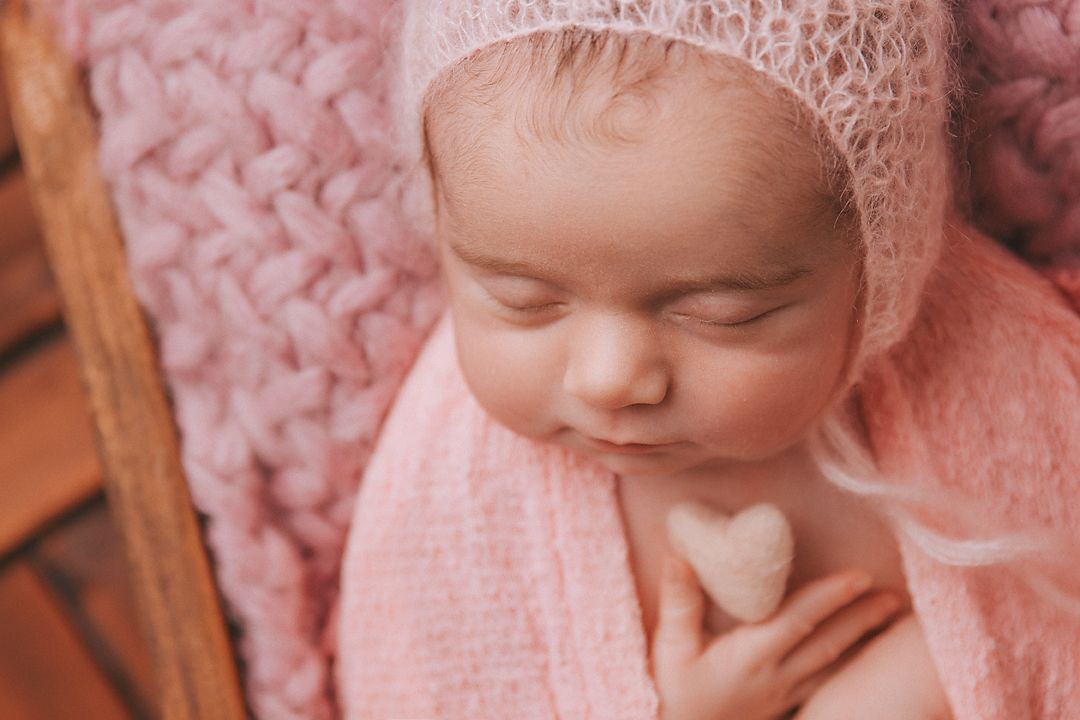 Baby wearing white lace bonnet rests peacefully on pink textured blanket in soft natural lighting.