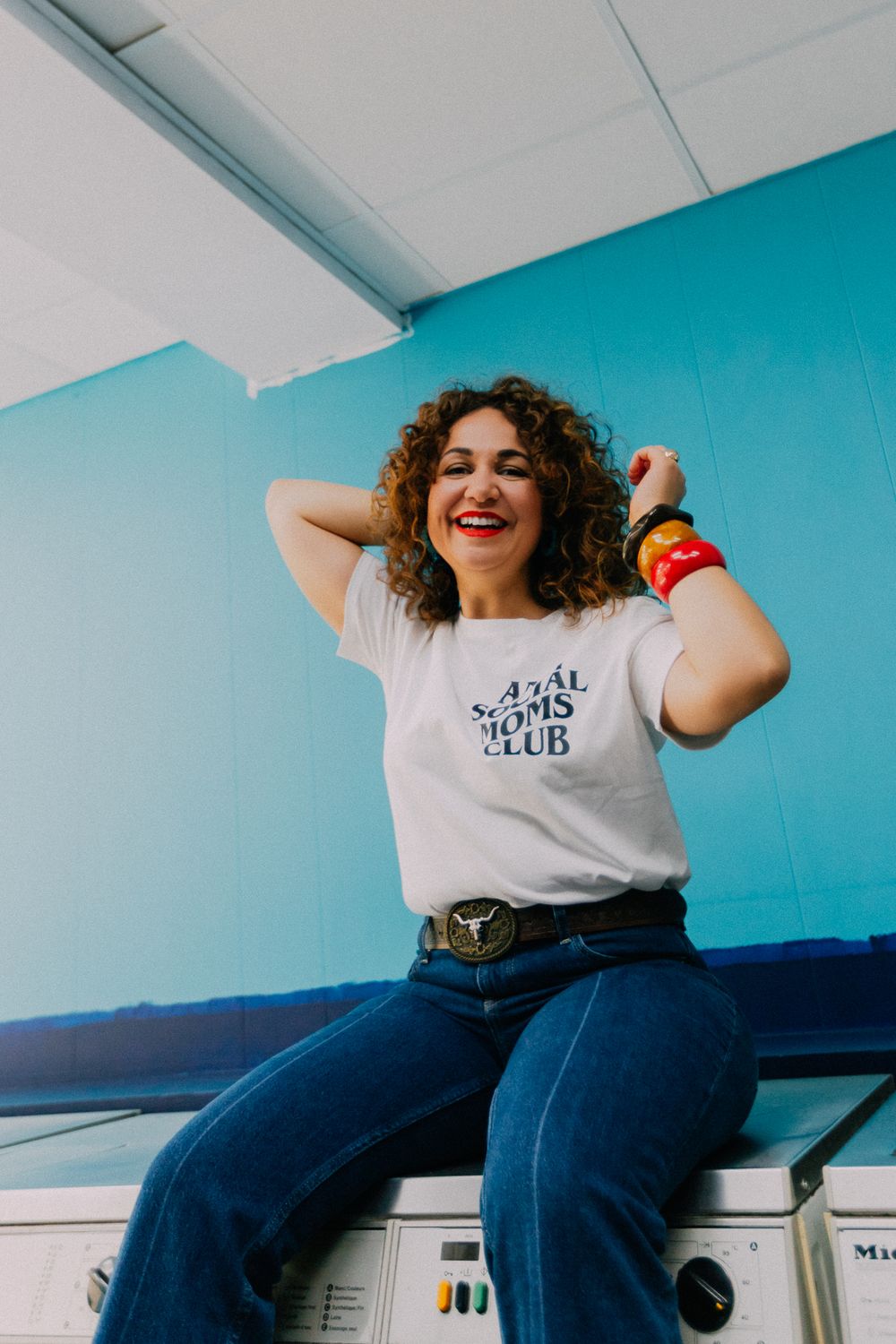 Laughing woman in white graphic tee and flare jeans sits atop washing machines in a bright blue laundromat.