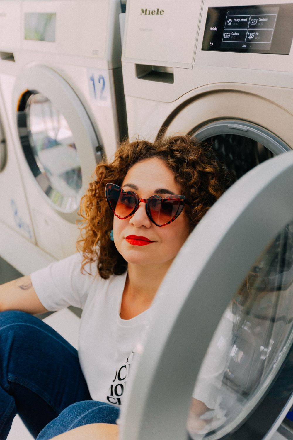 Woman with curly hair and red sunglasses poses by Miele washing machines in a laundromat.