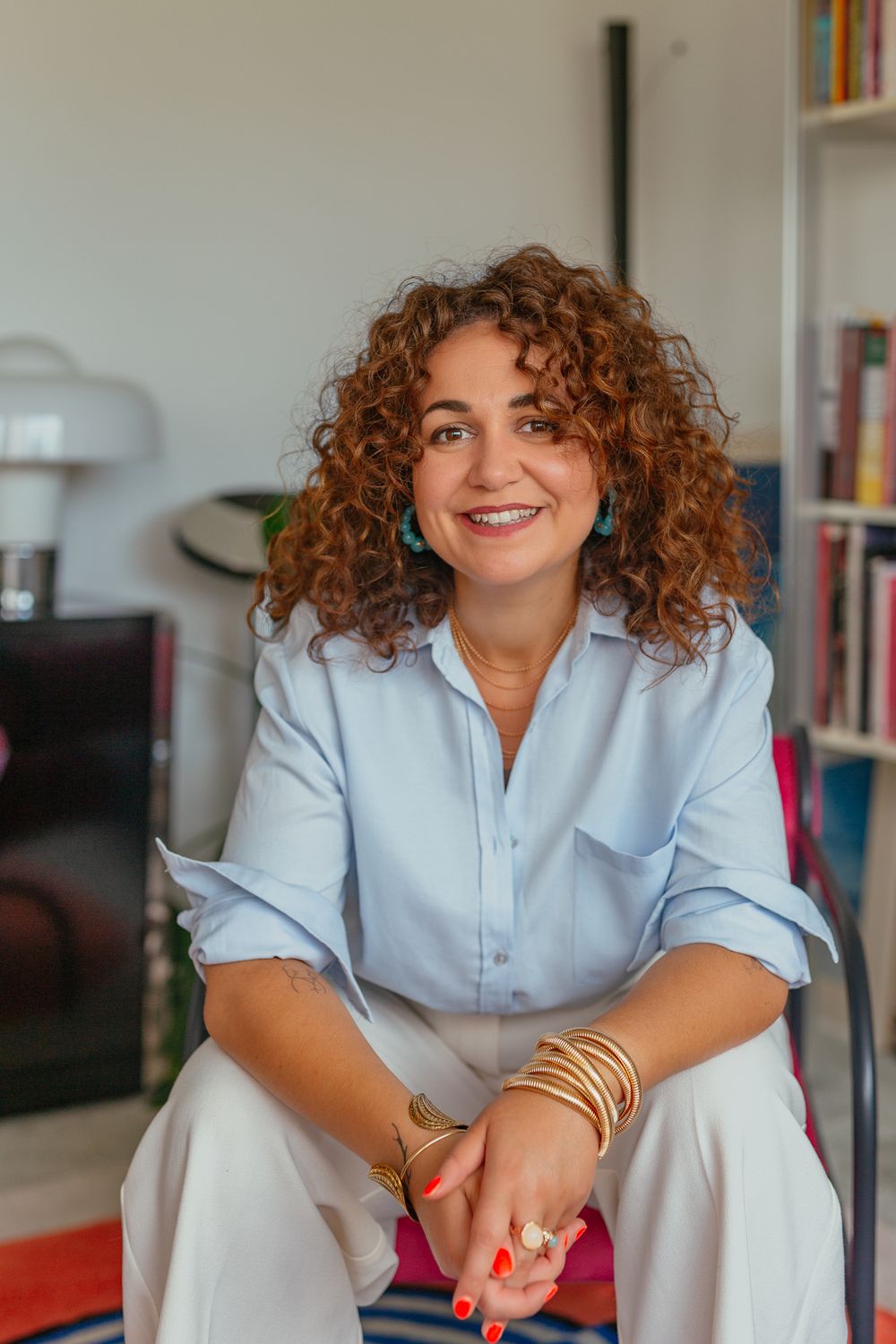 Smiling curly-haired woman in light blue shirt and white pants seated in bright room with bookshelf background.