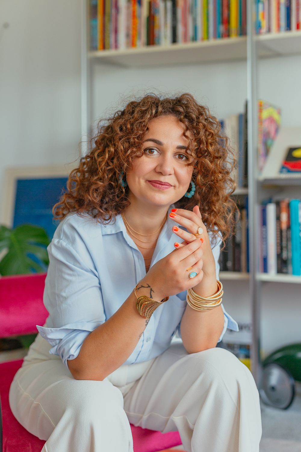 Curly-haired woman in blue shirt and white pants holding phone, seated in front of colorful bookshelf.
