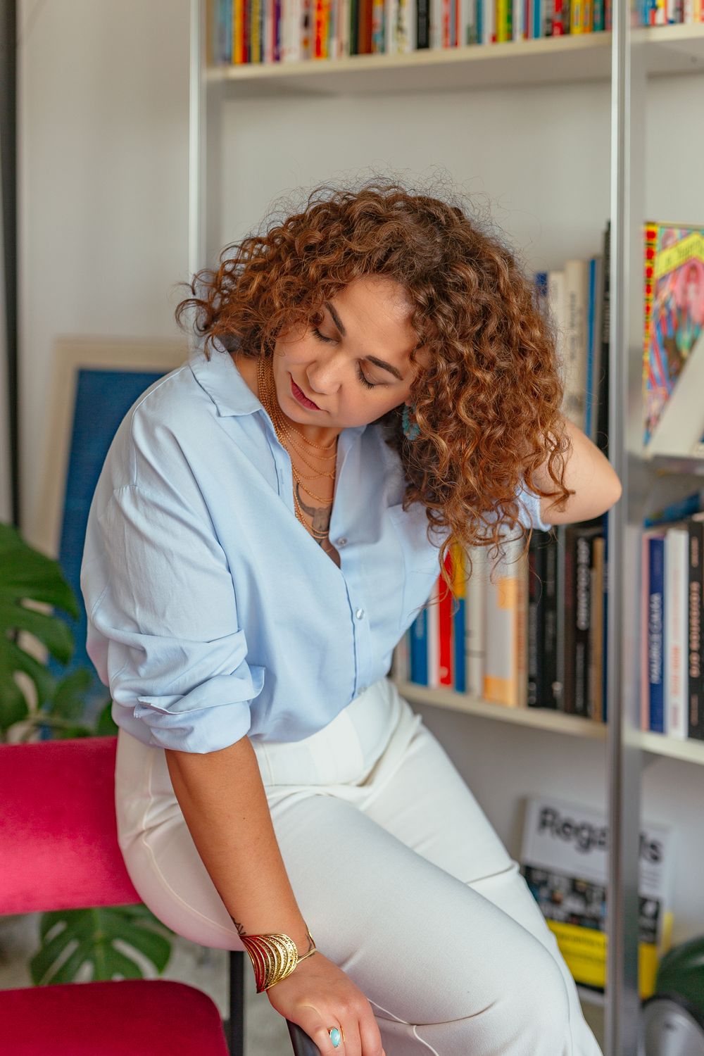 Curly-haired woman in blue shirt and white pants leaning against bookshelf, wearing gold bracelets.