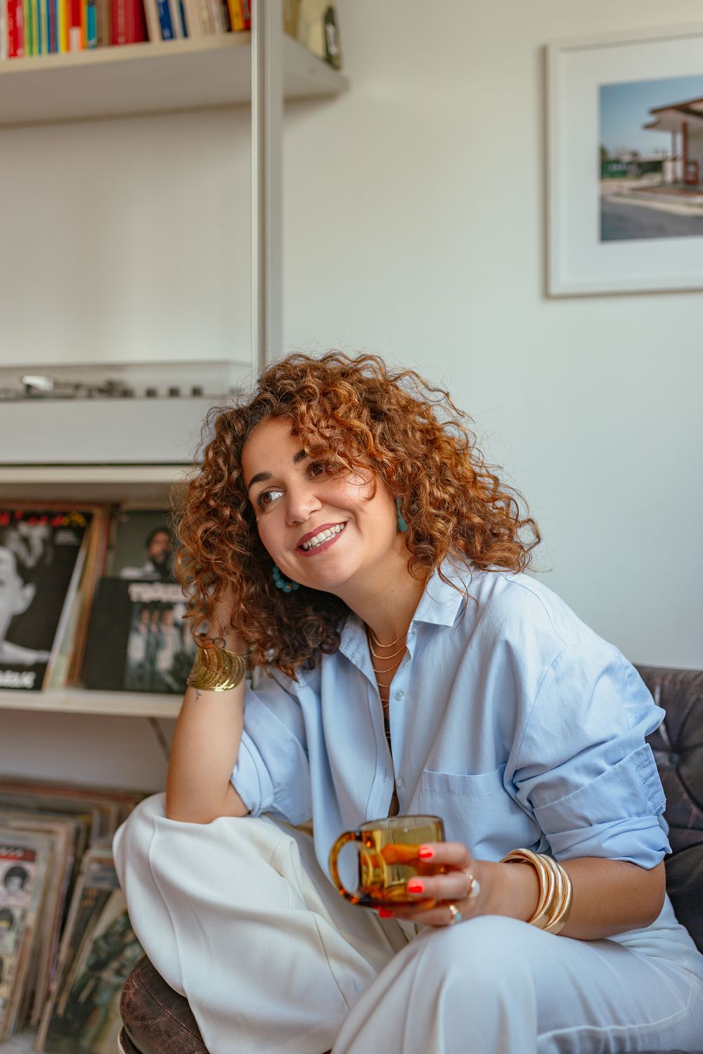 Smiling woman with curly red hair holding amber glass cup, wearing light blue shirt and gold bracelets, sitting by vinyl record shelf.
