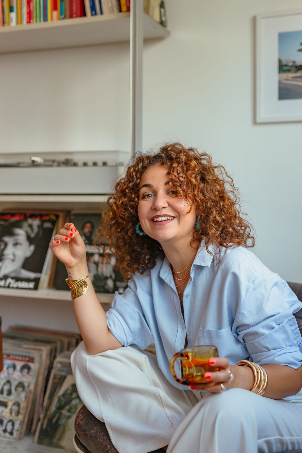 Happy curly-haired woman in blue shirt holding tea glass, wearing turquoise earrings and gold bracelets near vinyl record collection.
