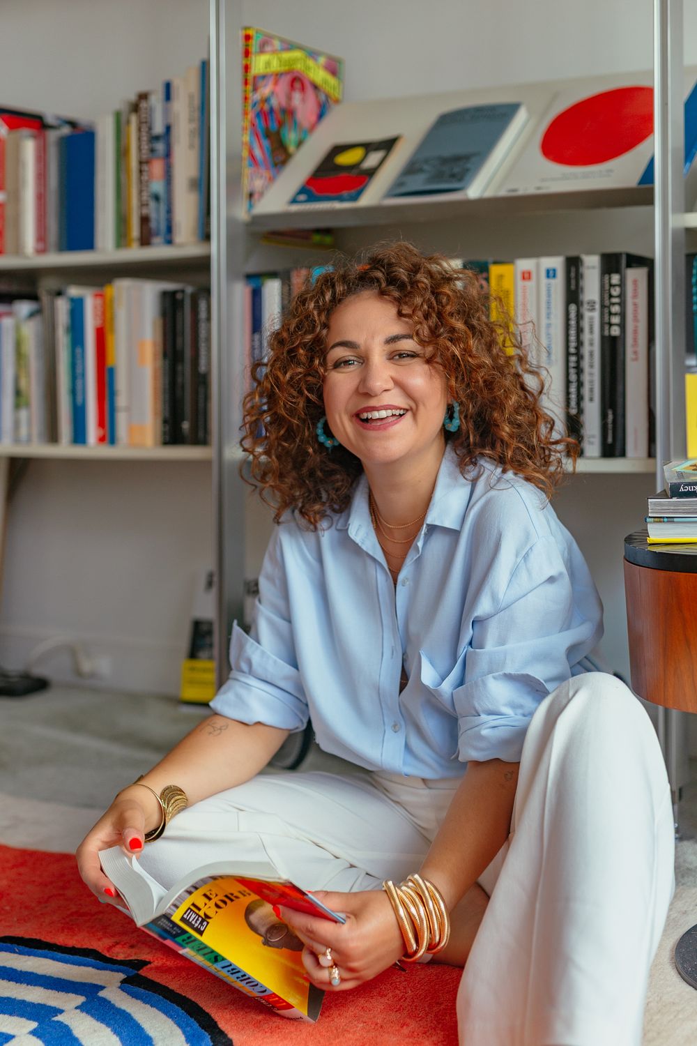 Cheerful woman with curly hair sitting on floor holding magazine, wearing blue shirt and gold bangles in front of bookshelf.