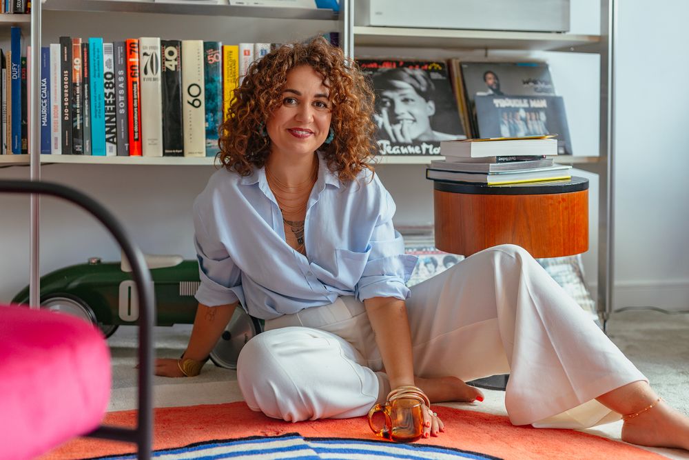 Relaxed woman with curly auburn hair in blue shirt and white pants sitting on orange rug near colorful bookshelf.