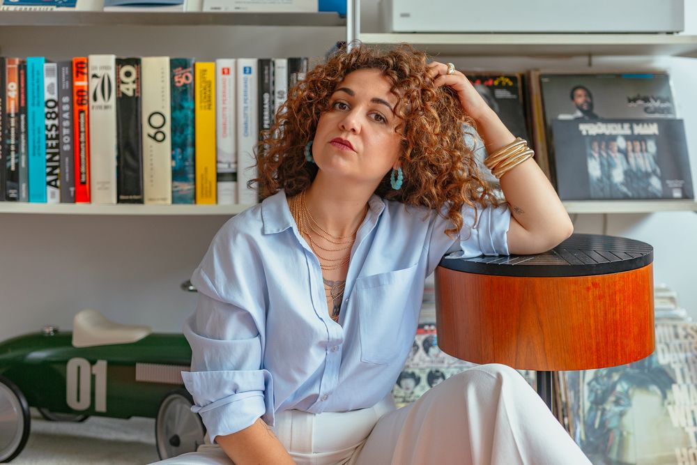 Pensive curly-haired woman in light blue shirt leaning on wooden lamp, sitting beside bookshelf with vintage toy car.