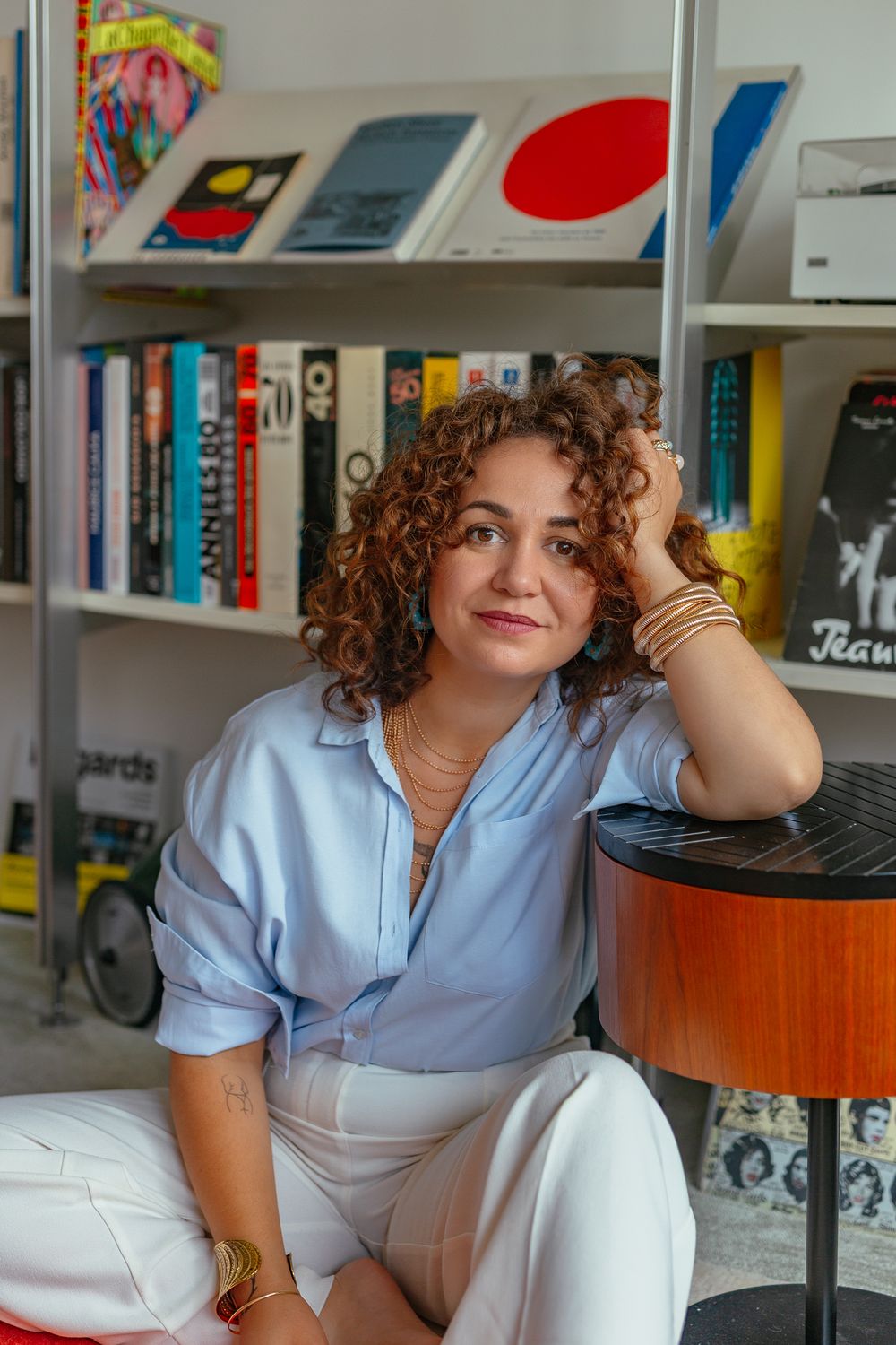 Woman with curly hair resting head on hand, wearing blue shirt and gold bracelets, seated beside eclectic bookshelf.