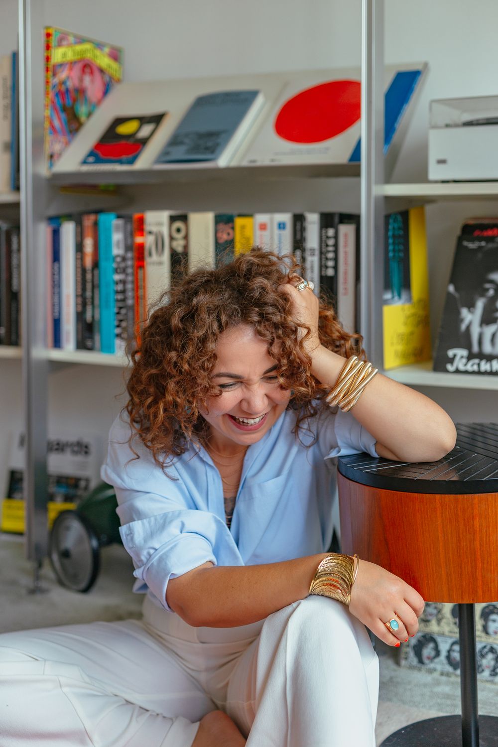 Laughing woman with curly auburn hair in blue shirt and white pants, wearing gold cuffs, sitting by colorful bookshelf.
