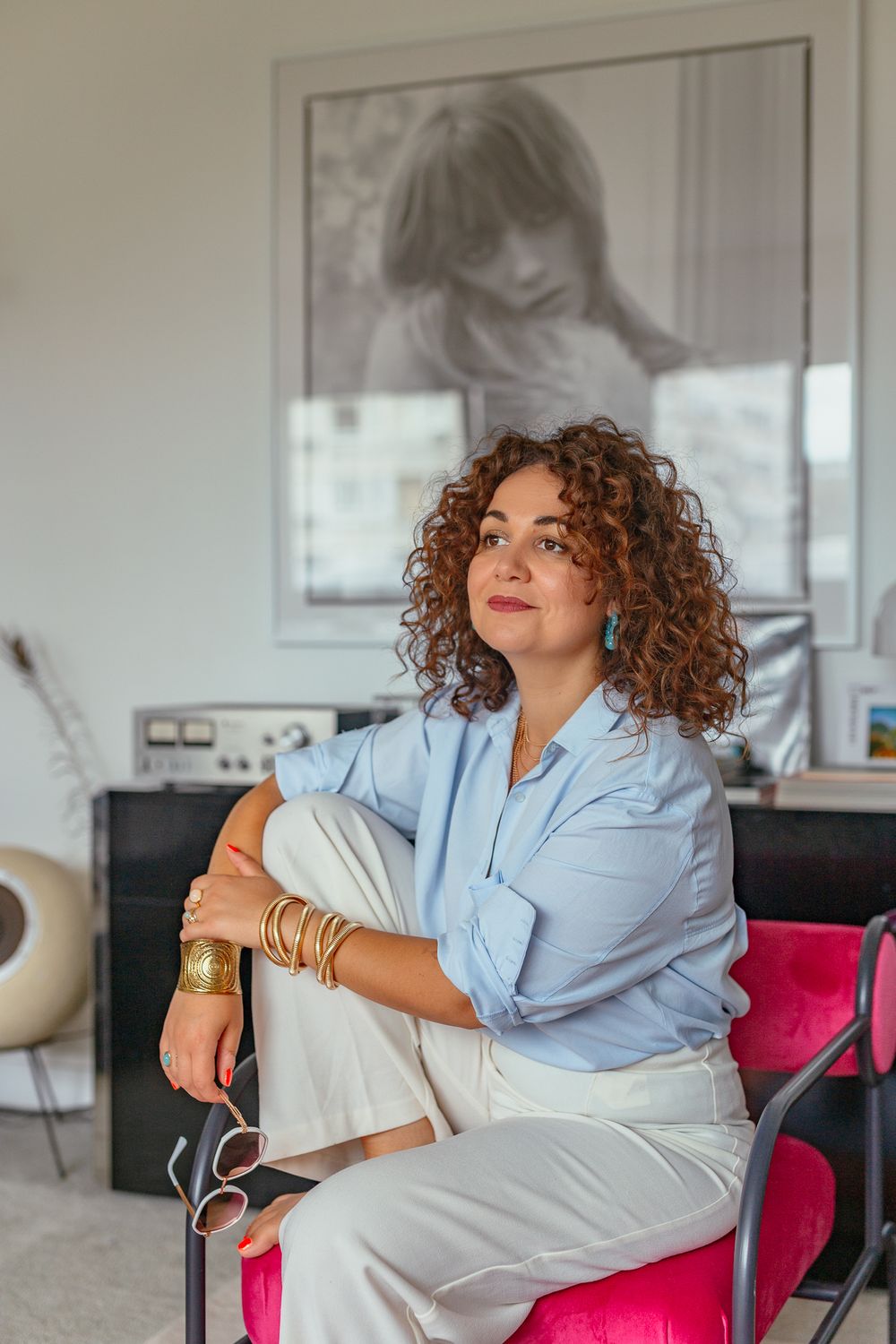 Thoughtful curly-haired woman in blue shirt and white pants sitting in pink chair beneath large black and white portrait.