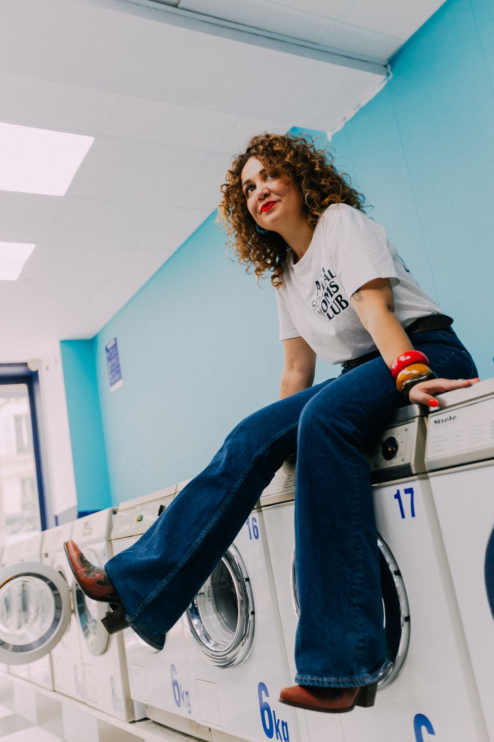 Stylish curly-haired woman sitting atop washing machines in laundromat, wearing white graphic tee and wide-leg denim jeans.