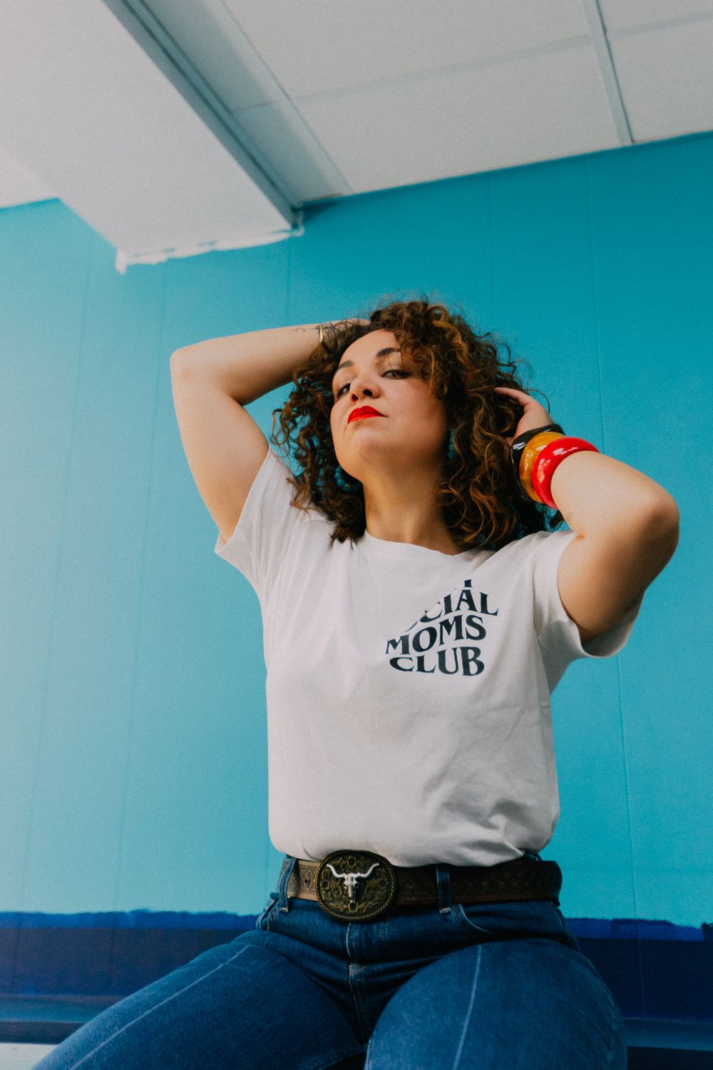 Woman with curly hair wearing Social Moms Club white tee and denim jeans posing against bright turquoise blue wall.