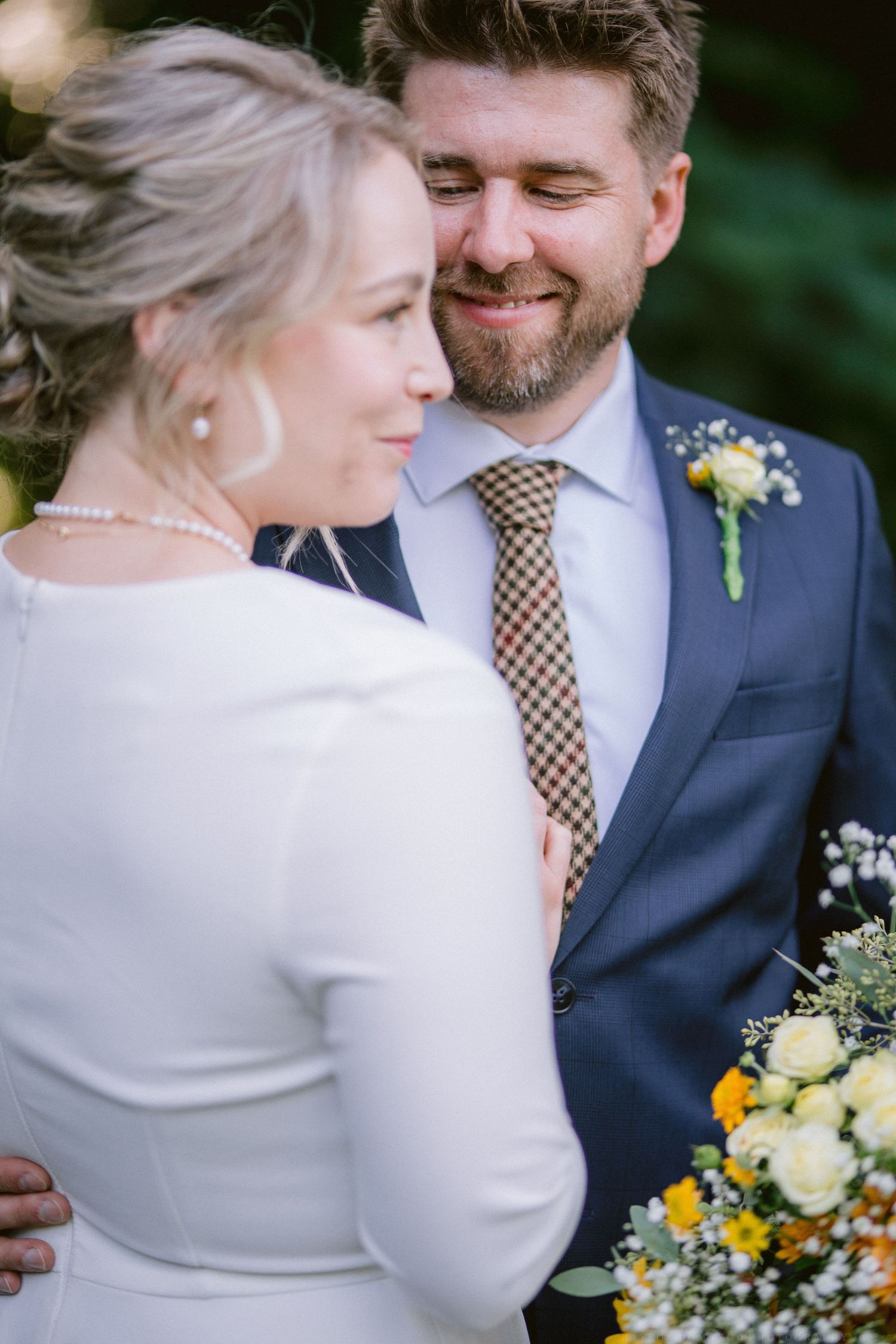 Wedding couple shares an intimate moment in formal attire with yellow and white floral bouquet.