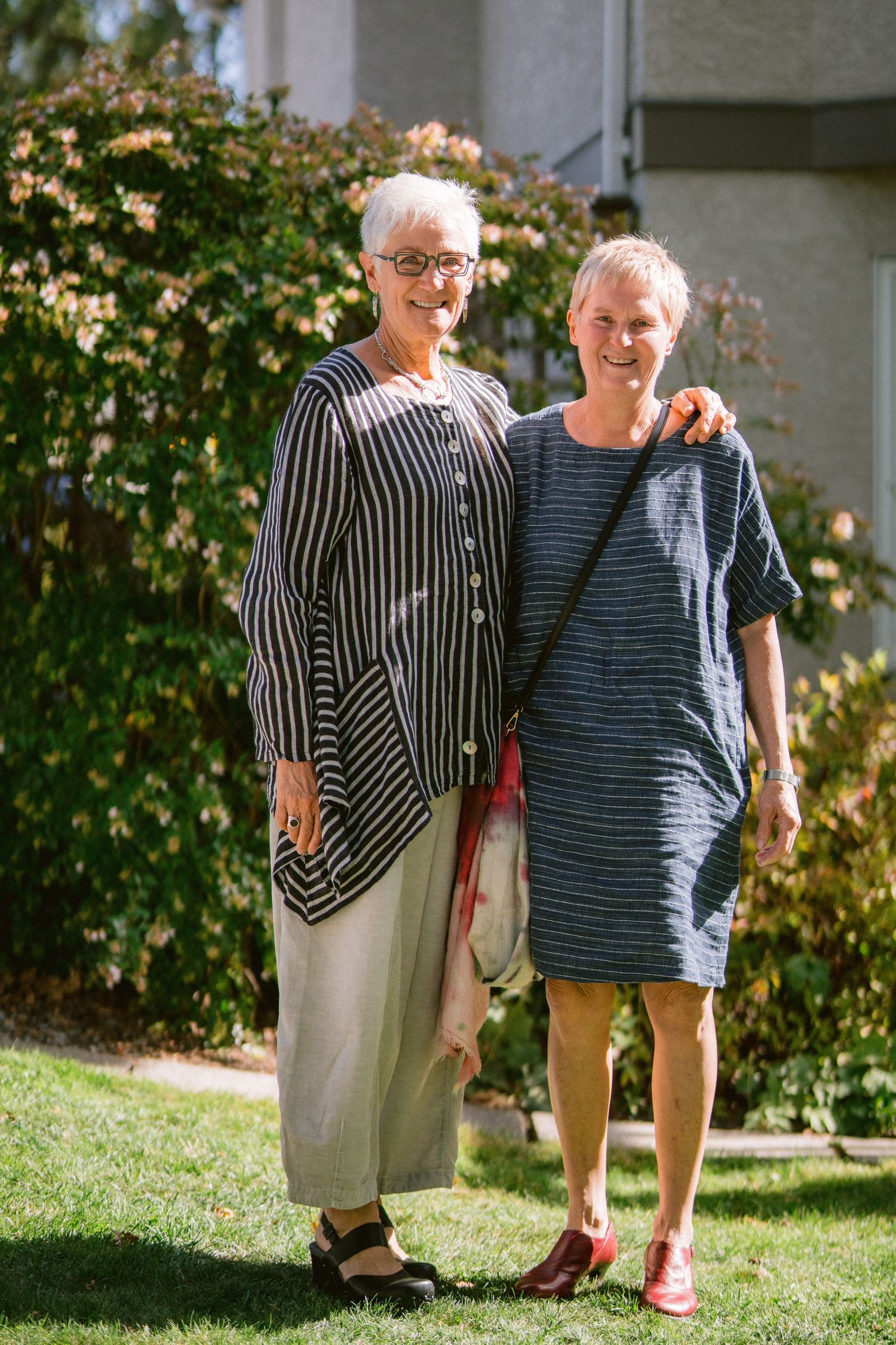 Two people in casual attire share a warm moment together on a sunny lawn with blooming flowers in the background.