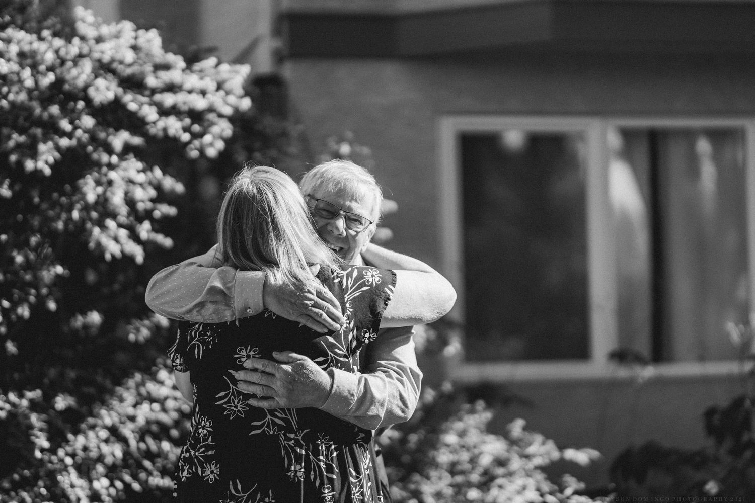 Black and white image showing people embracing in a series of emotional reunion moments with blooming flowers in background.