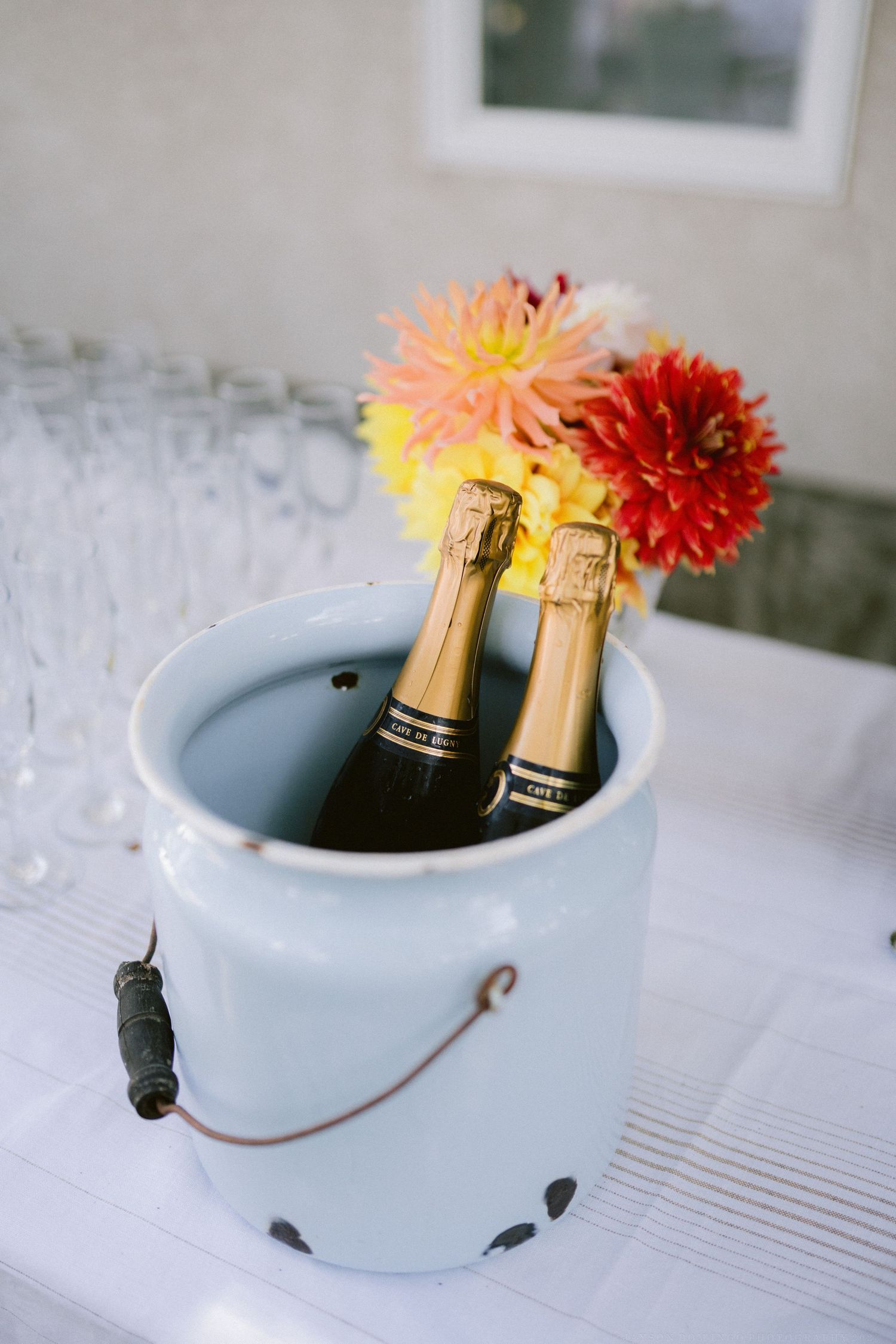 Champagne bottle and colorful dahlias arranged in a light blue ceramic vase on a white tablecloth.