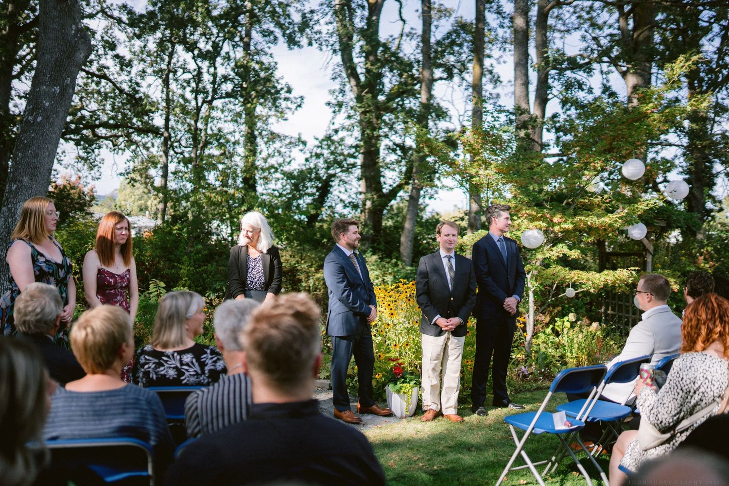 Outdoor wedding ceremony setting with guests gathered among trees in a natural woodland setting.