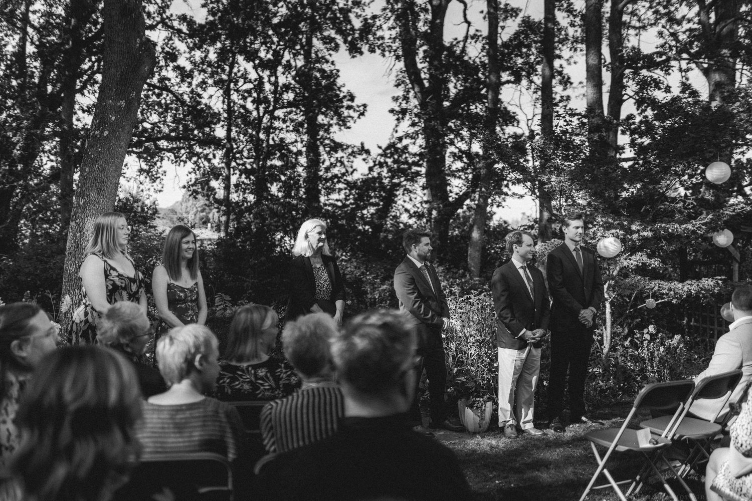 Black and white photograph of wedding guests gathered under trees at an outdoor ceremony.