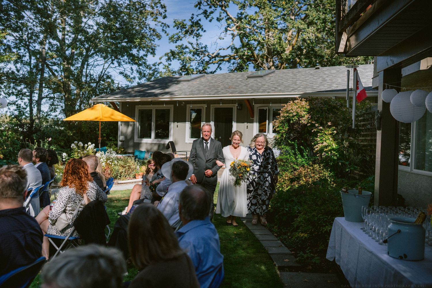 Outdoor wedding reception setup with guests mingling on a patio surrounded by trees.