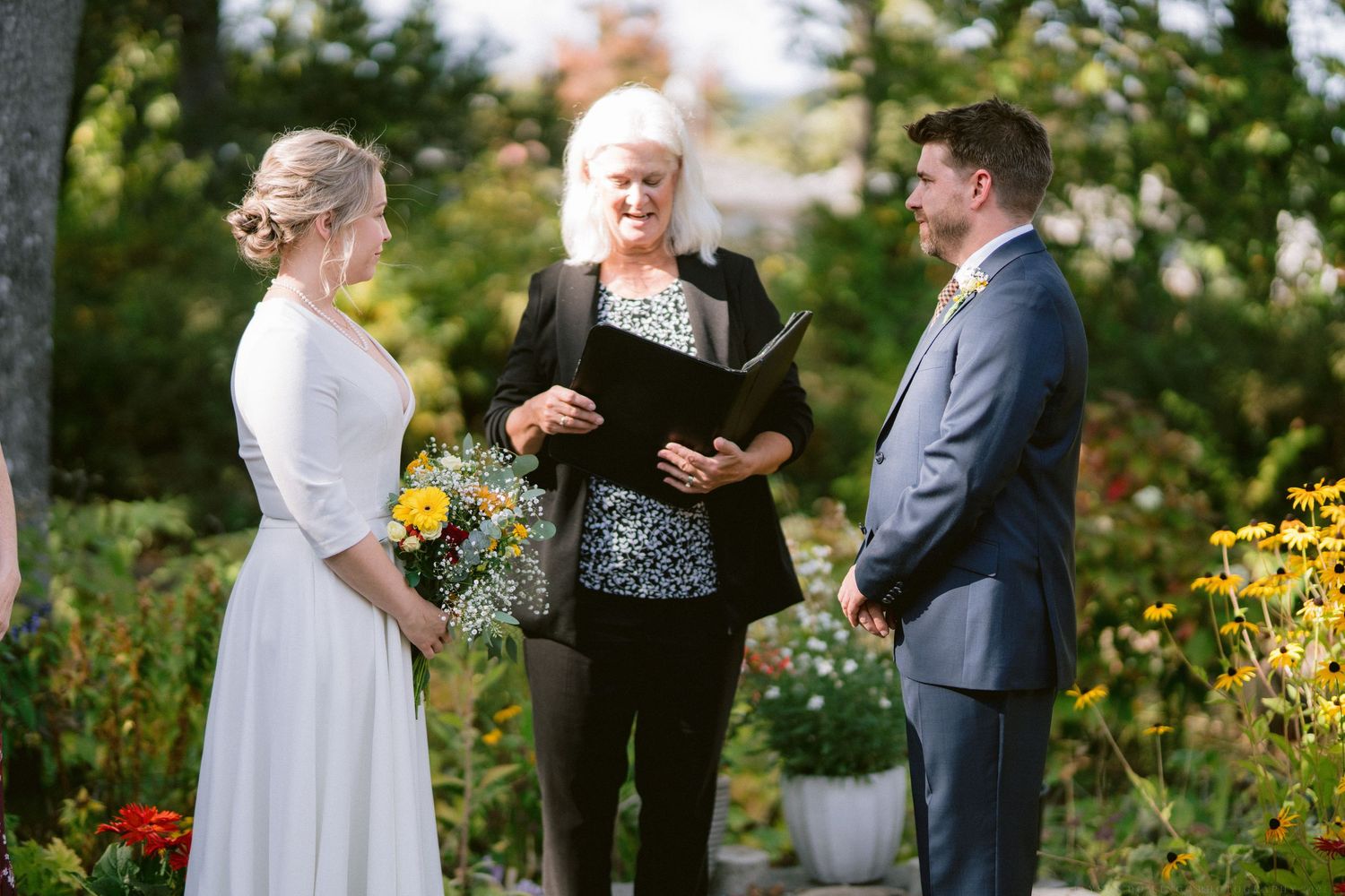 A wedding ceremony takes place in a lush garden setting with yellow flowers and greenery.