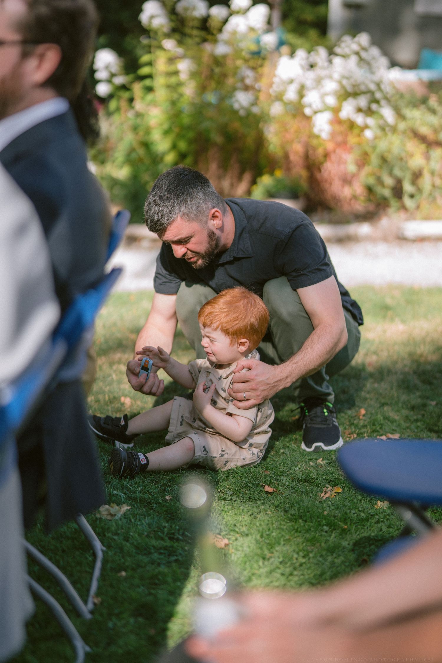 A tender moment shared between a man and small child with red hair sitting together on grass during an outdoor gathering.