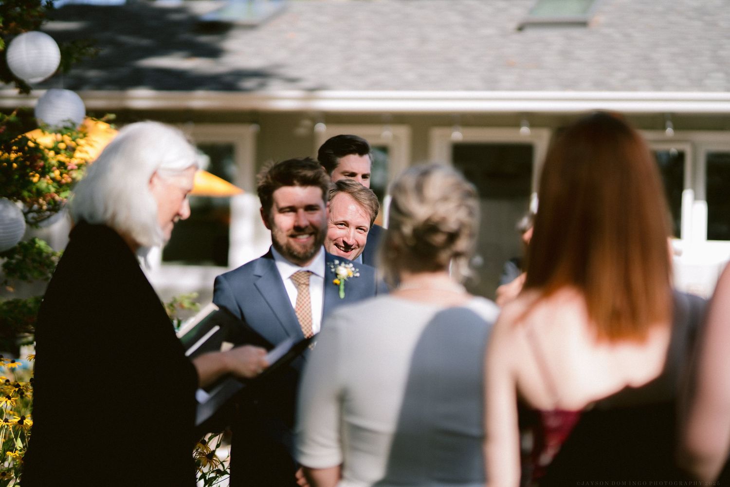 Wedding guests mingle during an outdoor ceremony with a white house visible in the background.