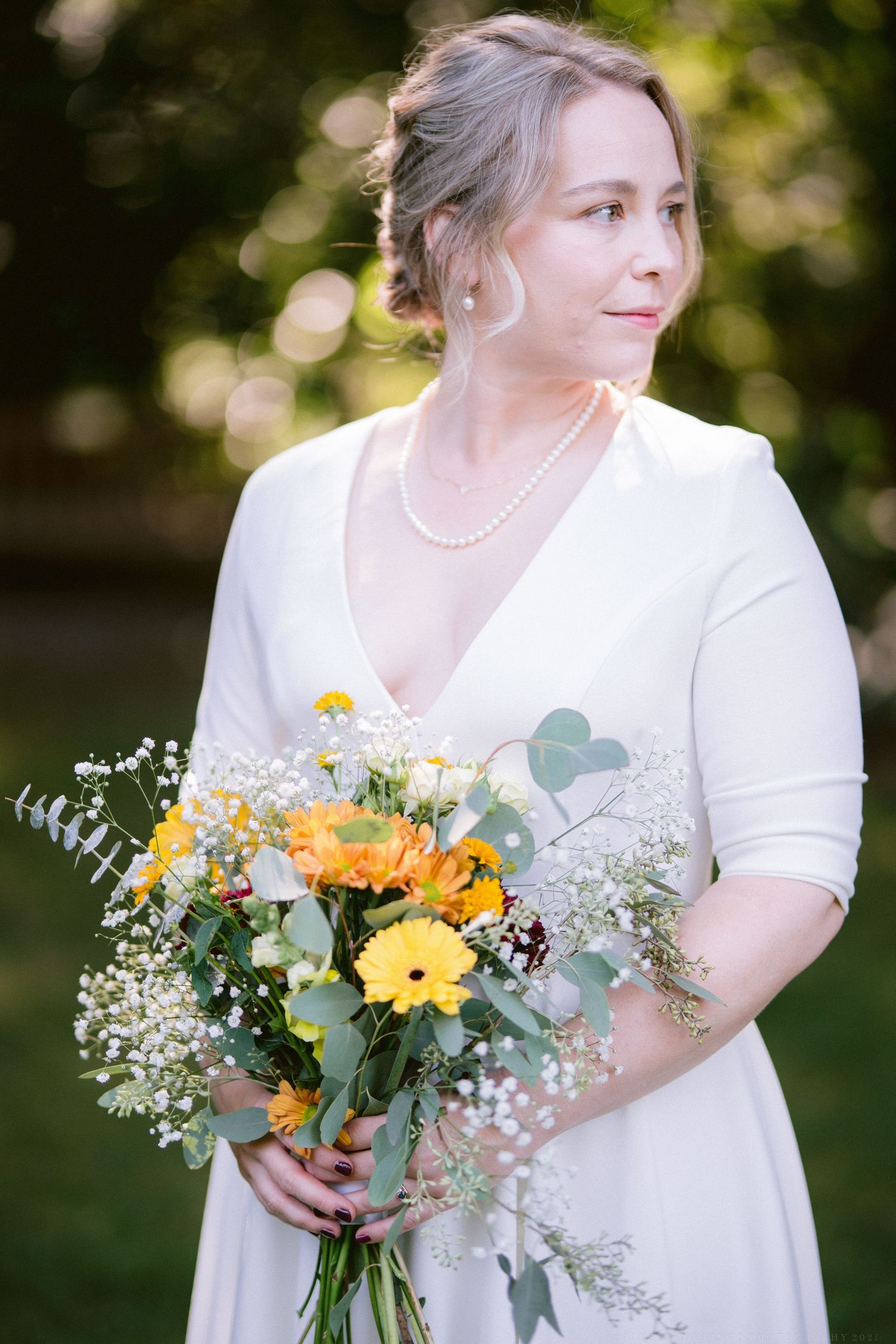 A bridal bouquet featuring vibrant yellow gerbera daisies, orange flowers, and delicate baby's breath against a blurred outdoor background.
