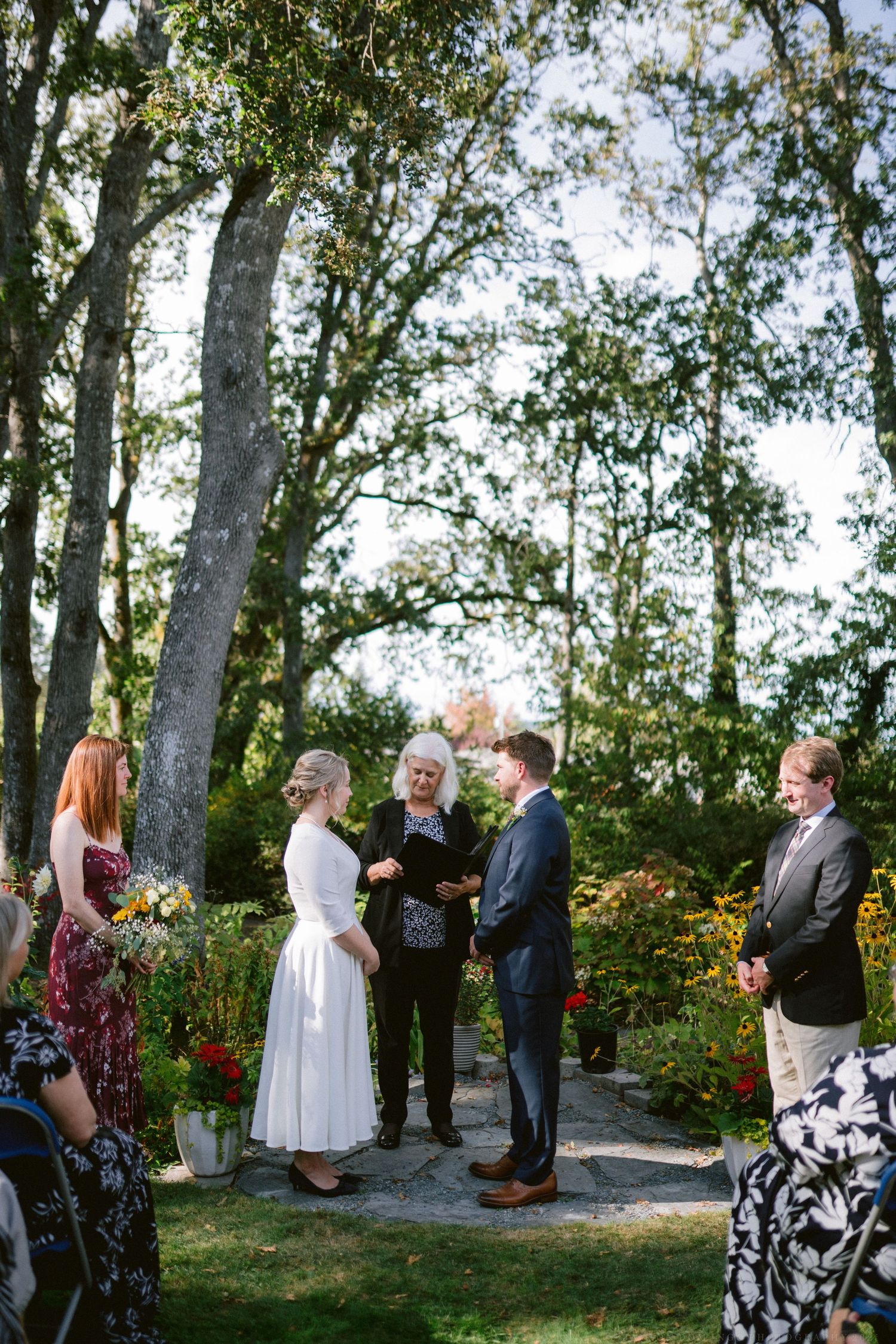 Wedding guests gathered outdoors among tall trees during a daytime ceremony.