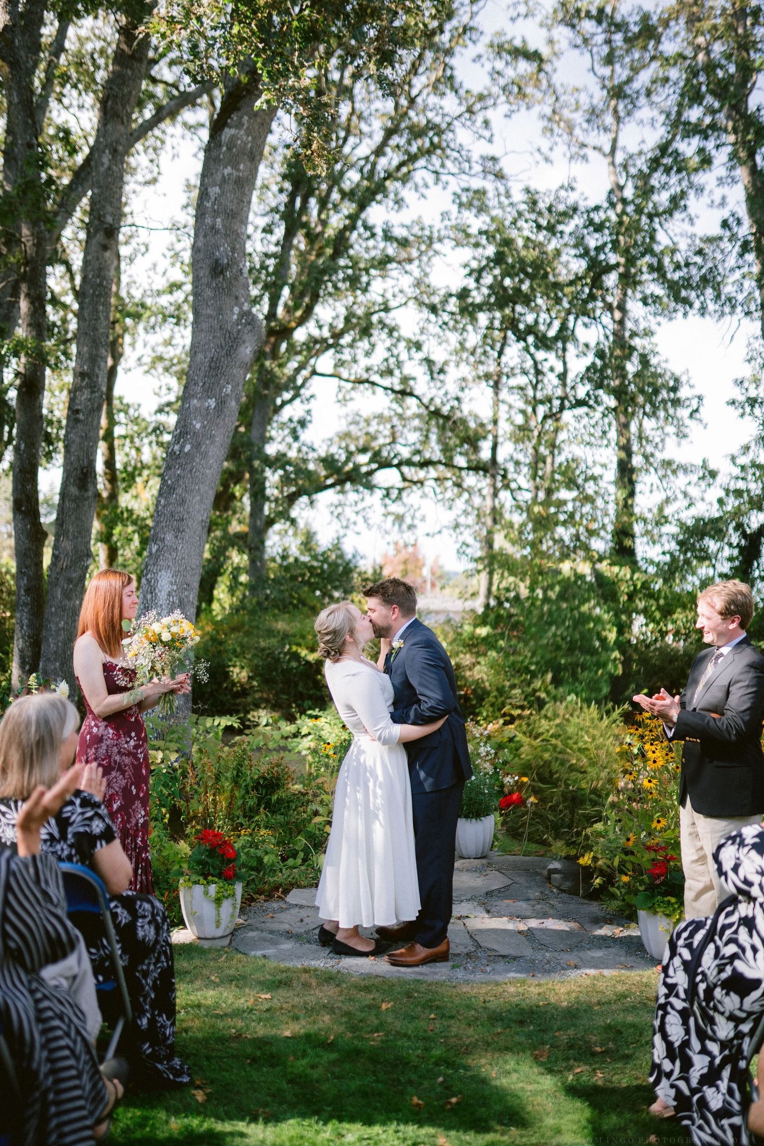 A wedding ceremony takes place outdoors among tall trees with guests gathered to witness the special moment.