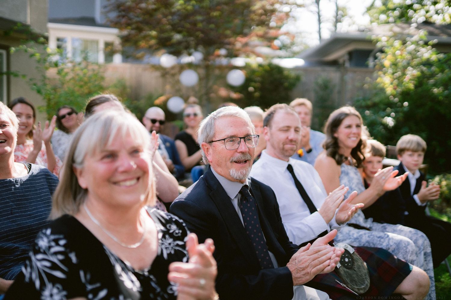 Wedding guests seated outdoors clapping and celebrating during a joyful ceremony moment.