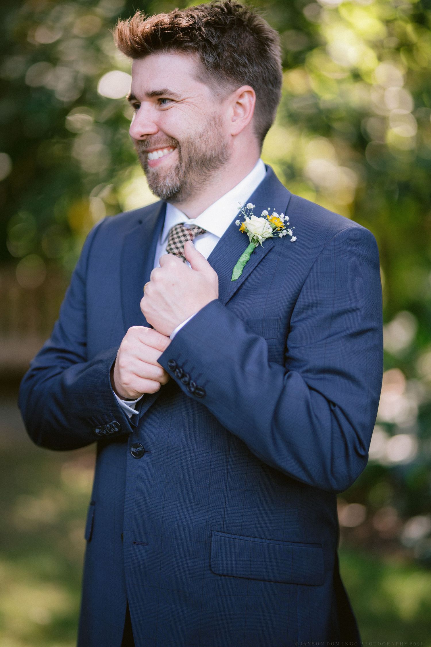 Groom in navy suit adjusting checkered tie and boutonniere in outdoor setting.
