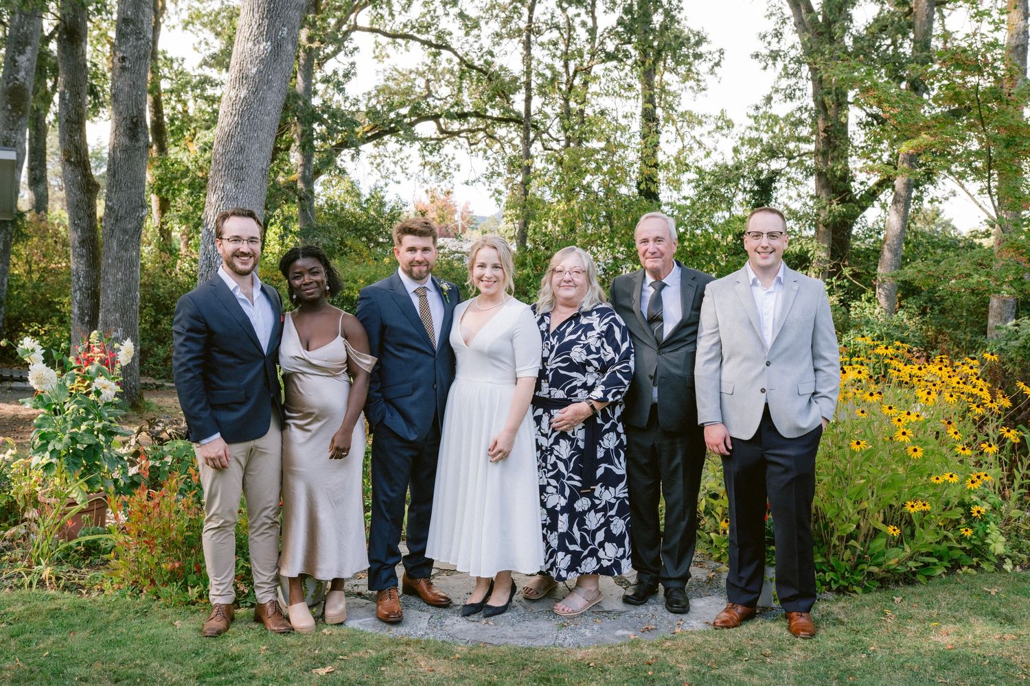 A group photo of wedding guests gathered outdoors in a garden setting with trees and orange flowers in the background.