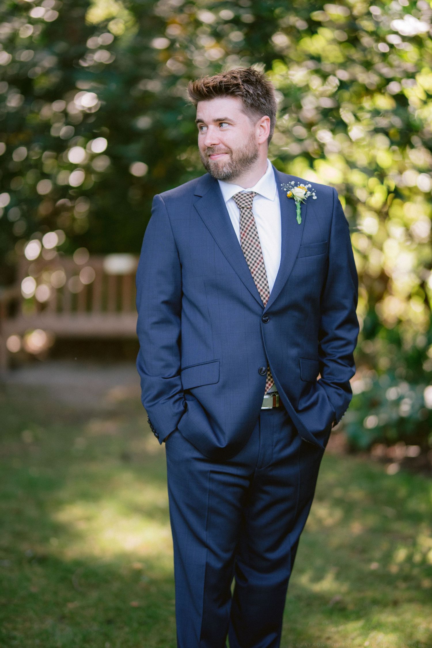 Groom in navy suit and polka dot tie poses in a sunlit garden setting.
