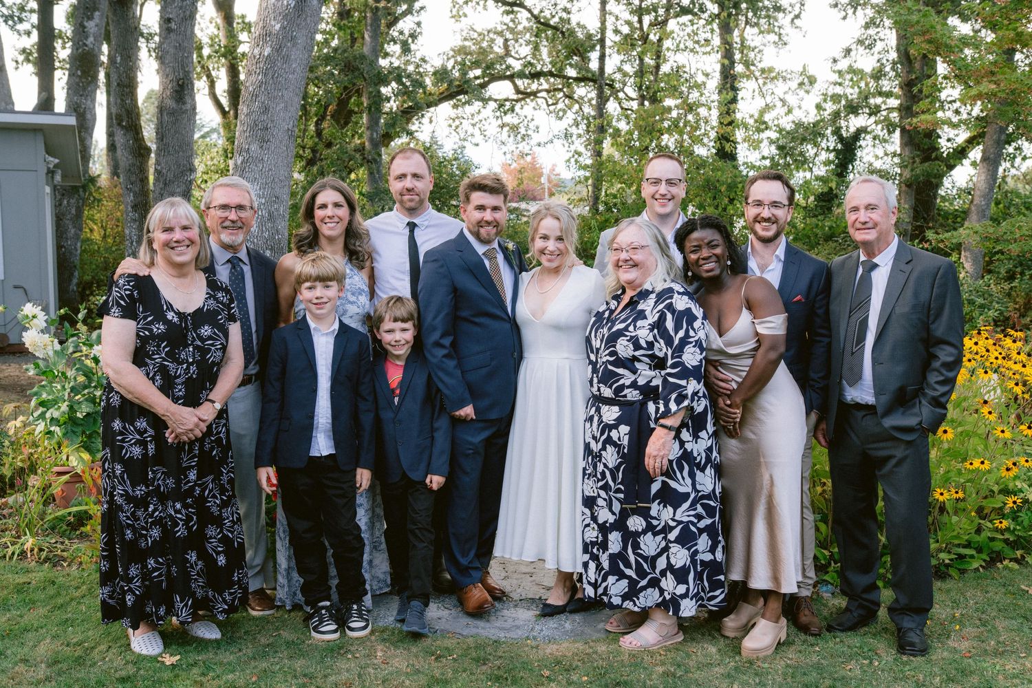 Wedding party and guests pose together in a garden setting with trees and colorful flowers visible behind them.