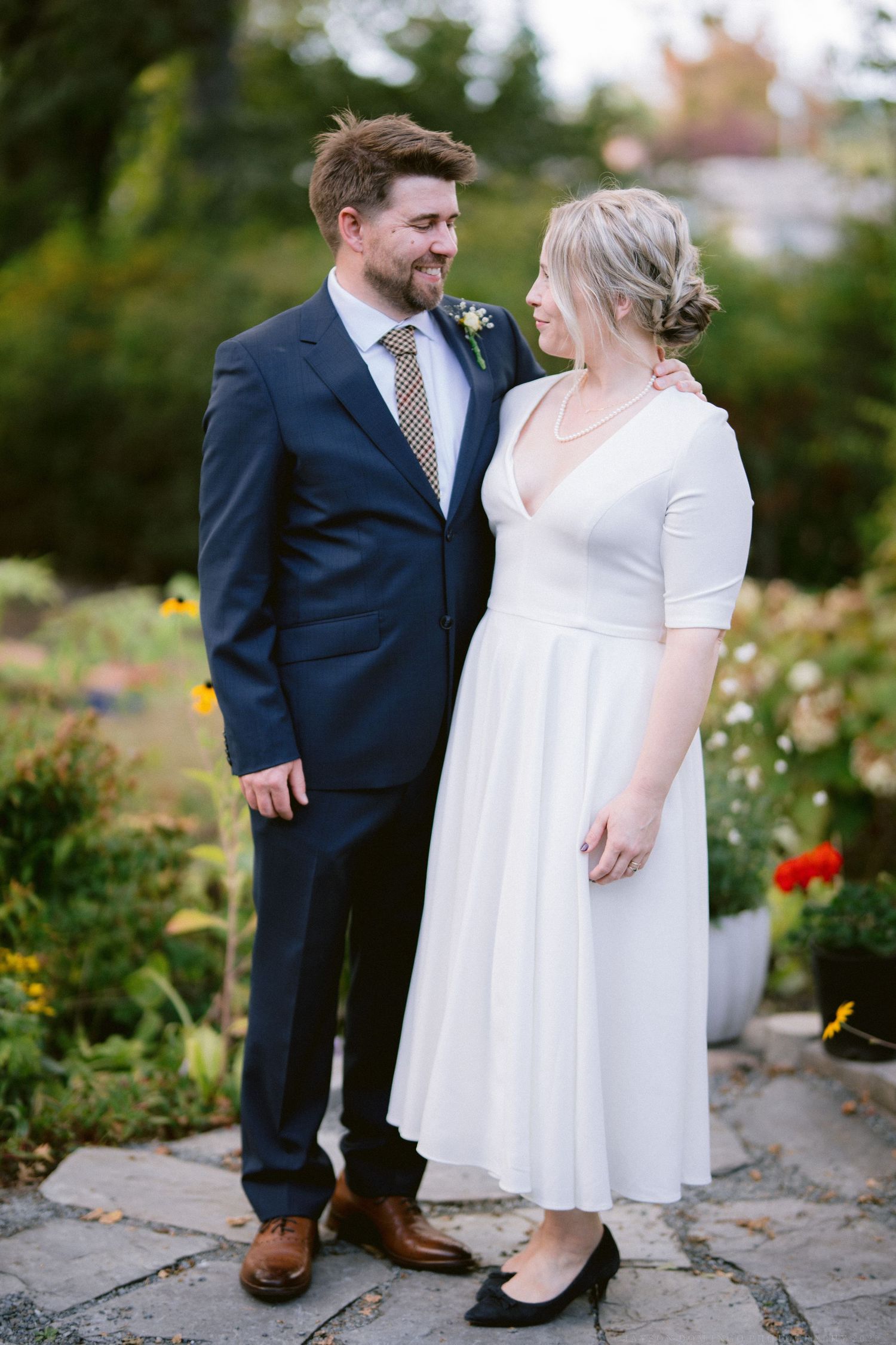 A romantic wedding photo shoot in a garden with the couple in formal attire standing among blooming flowers.