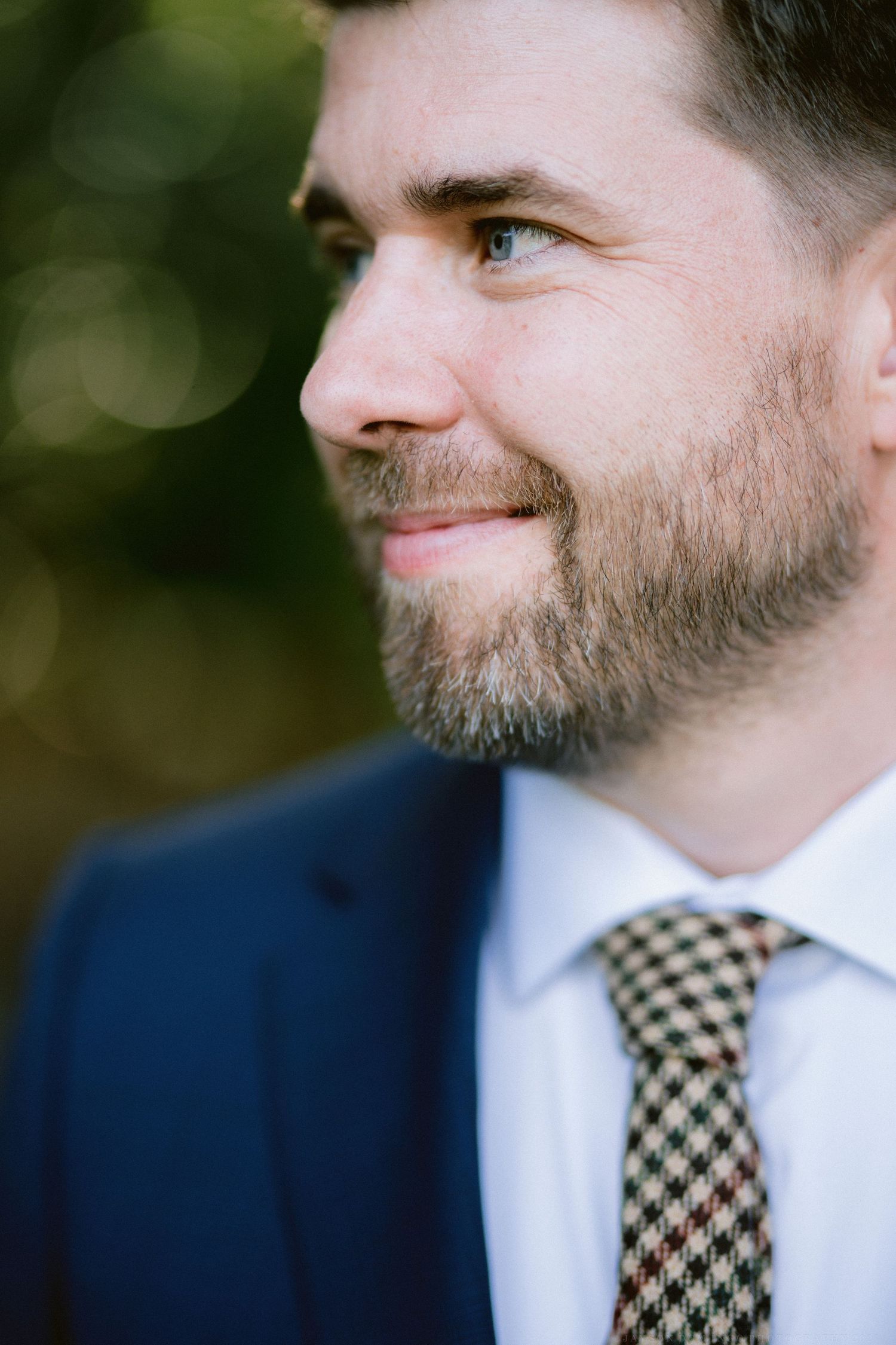 Close-up profile view of groom in navy suit with checkered tie and beard.