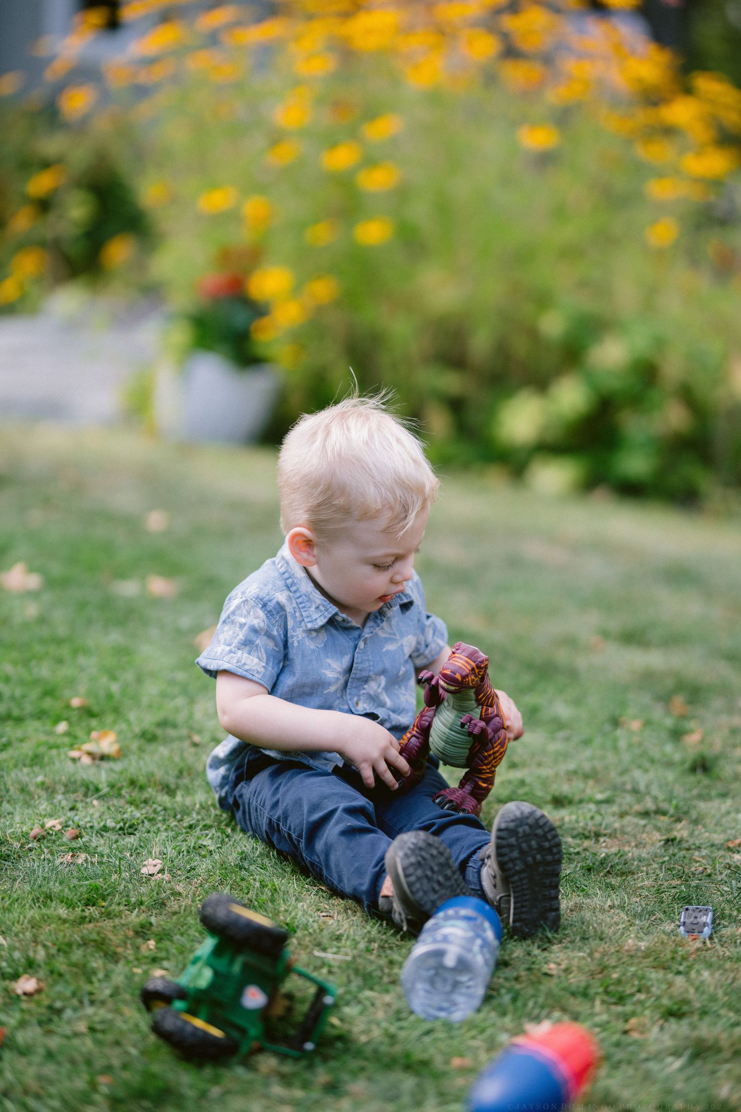 A toddler in denim overalls sits on grassy ground playing with toys among scattered yellow flowers in a garden.