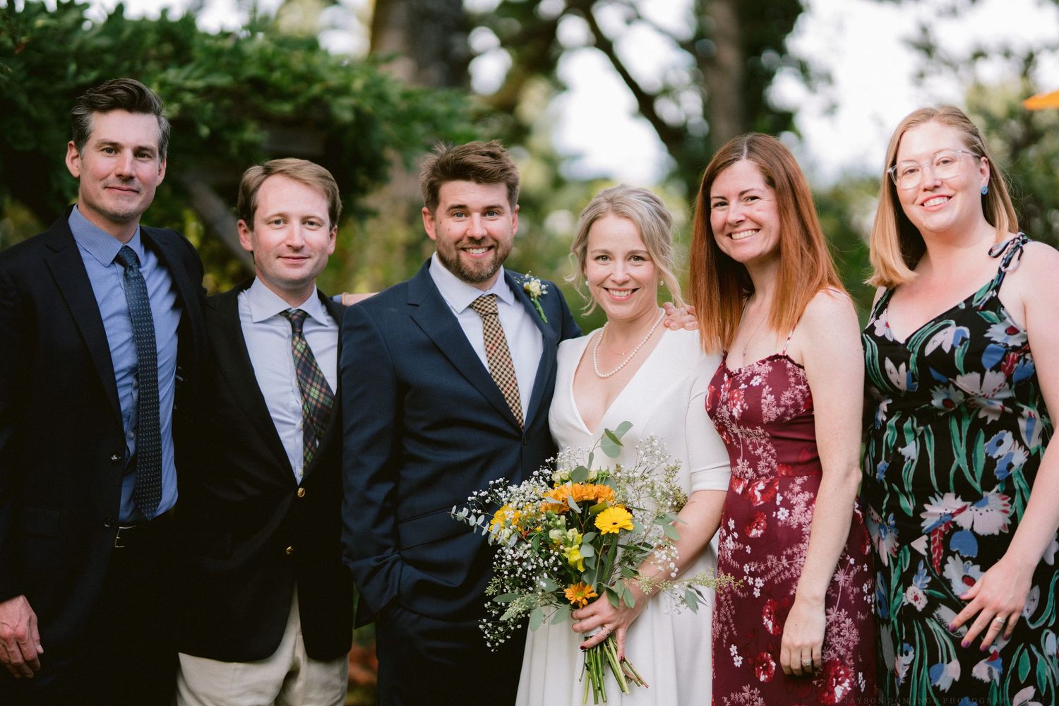 Wedding party poses outdoors in formal attire with burgundy bridesmaids dresses and yellow sunflower bouquets.