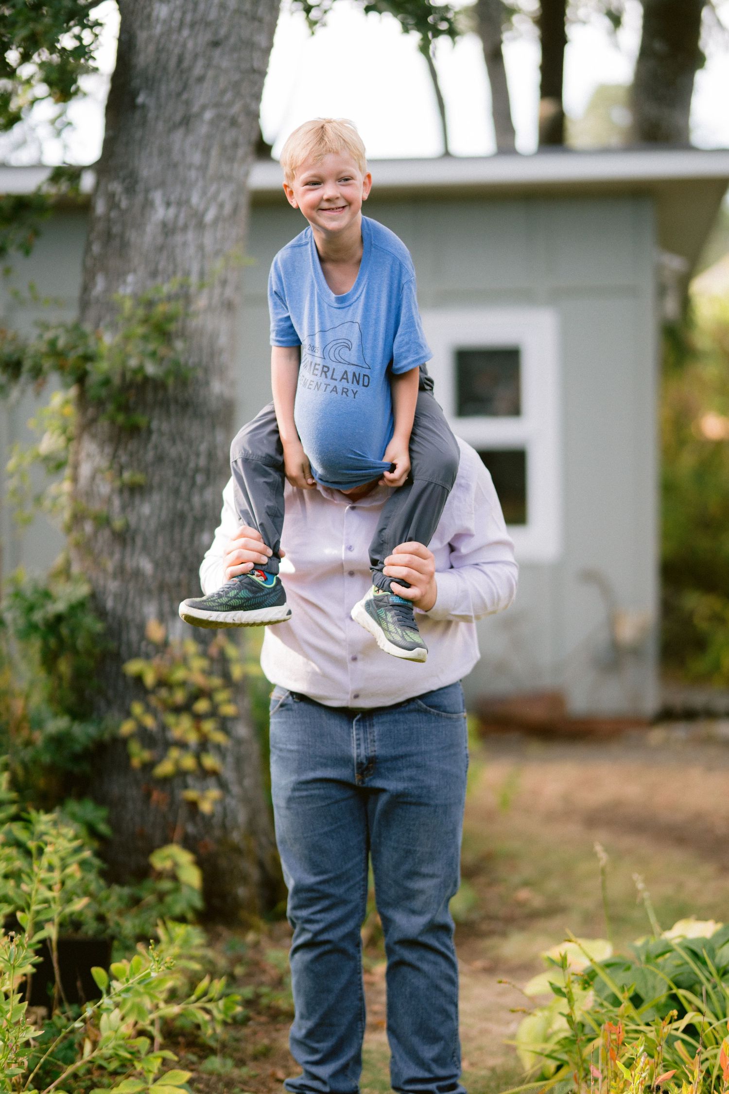 A child sits on someone's shoulders in a garden setting, wearing a blue t-shirt and smiling outdoors.