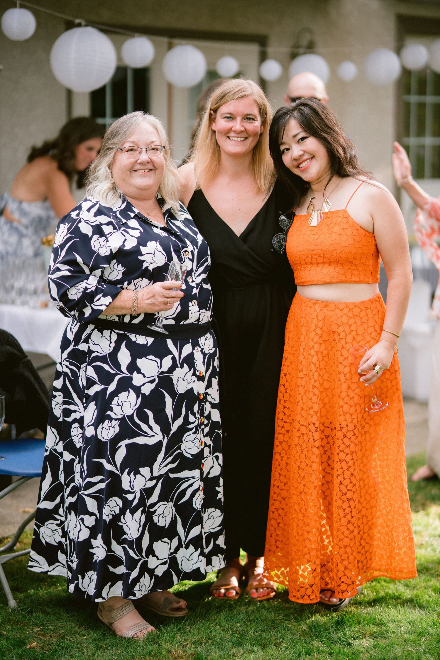 Wedding guests in floral and orange dresses sitting together on grass during an outdoor celebration.