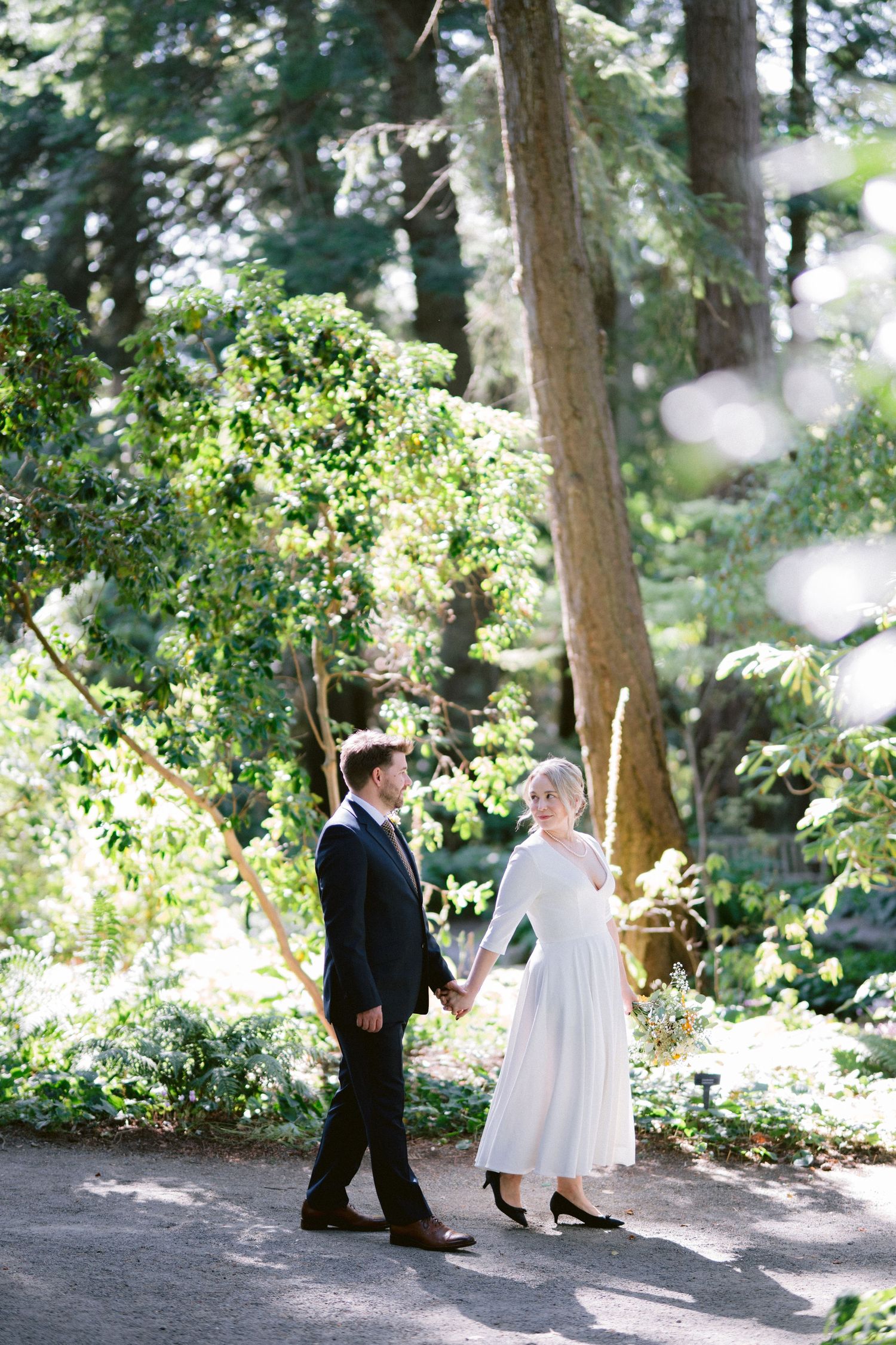 Wedding couple walks together on forest path surrounded by tall evergreen trees.