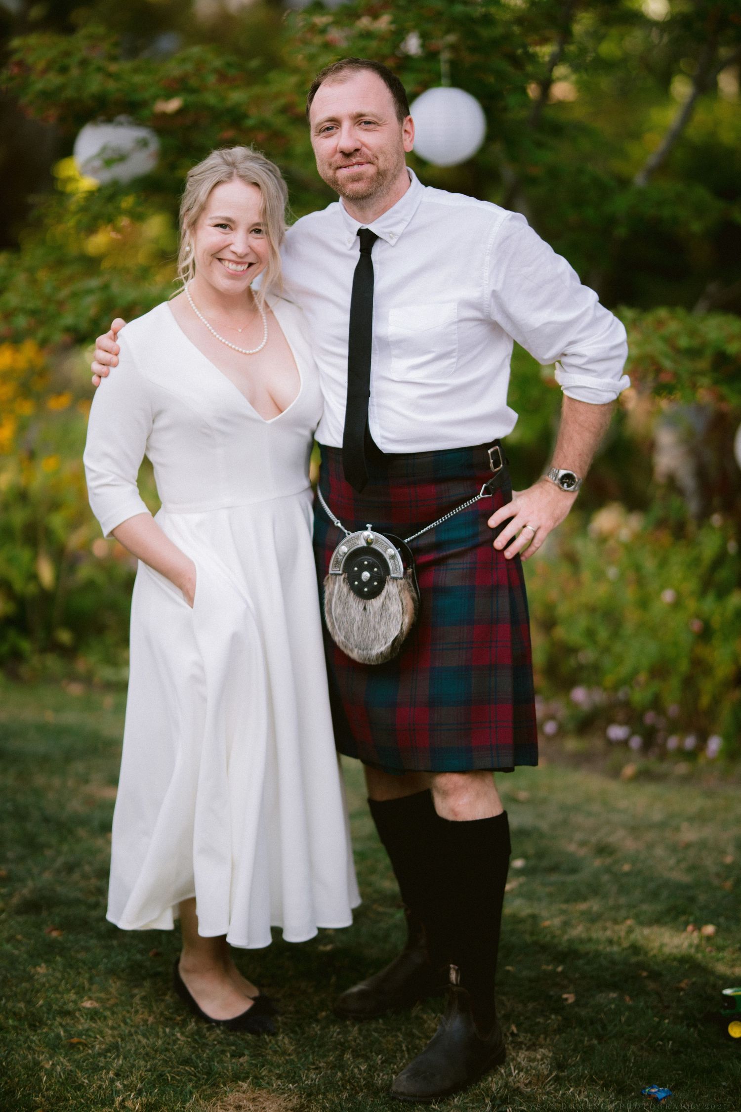A couple poses outdoors in wedding attire featuring a traditional Scottish kilt and sporran with a white dress.