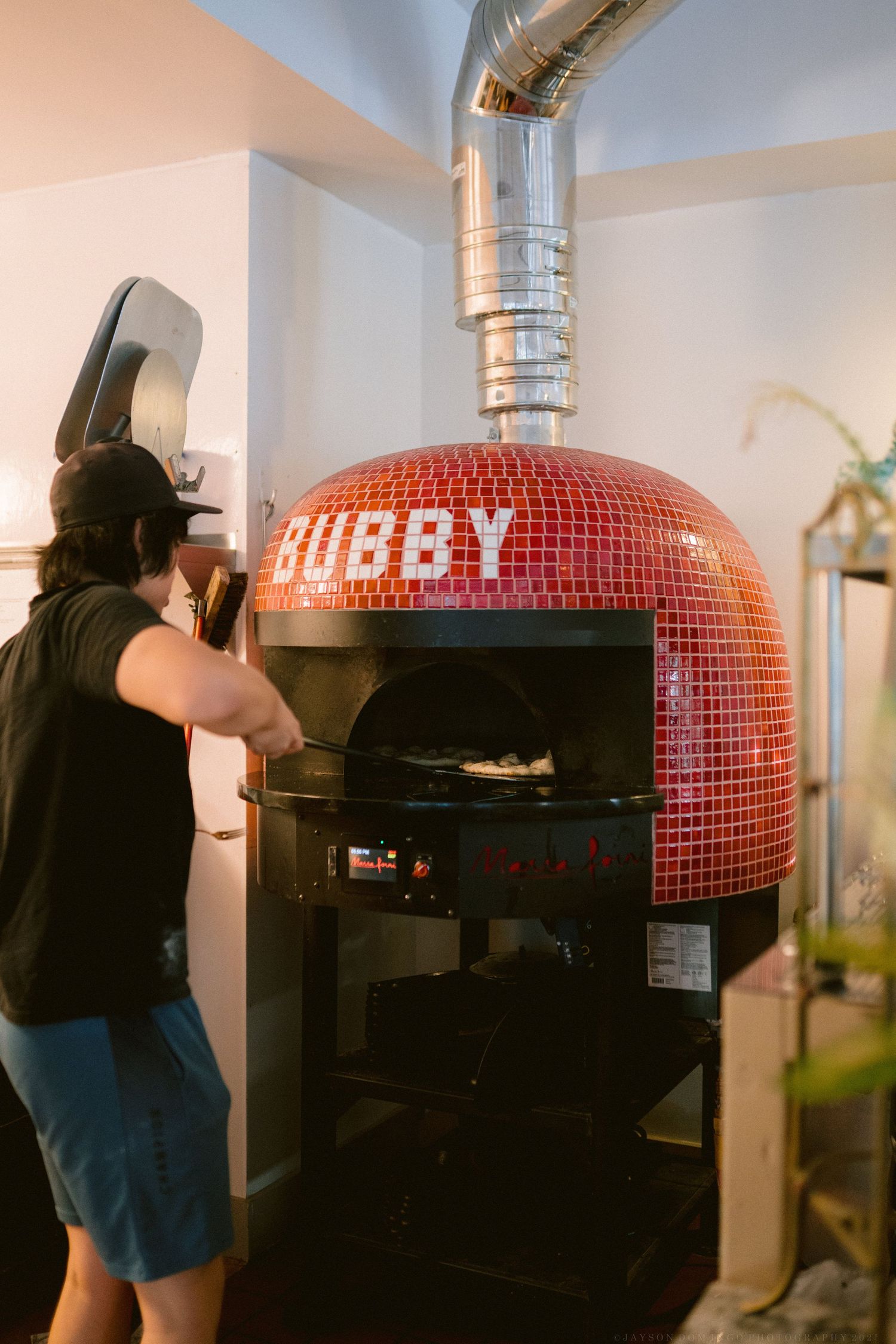 Chef preparing pizza in a traditional red-tiled wood-fired pizza oven in a restaurant kitchen.